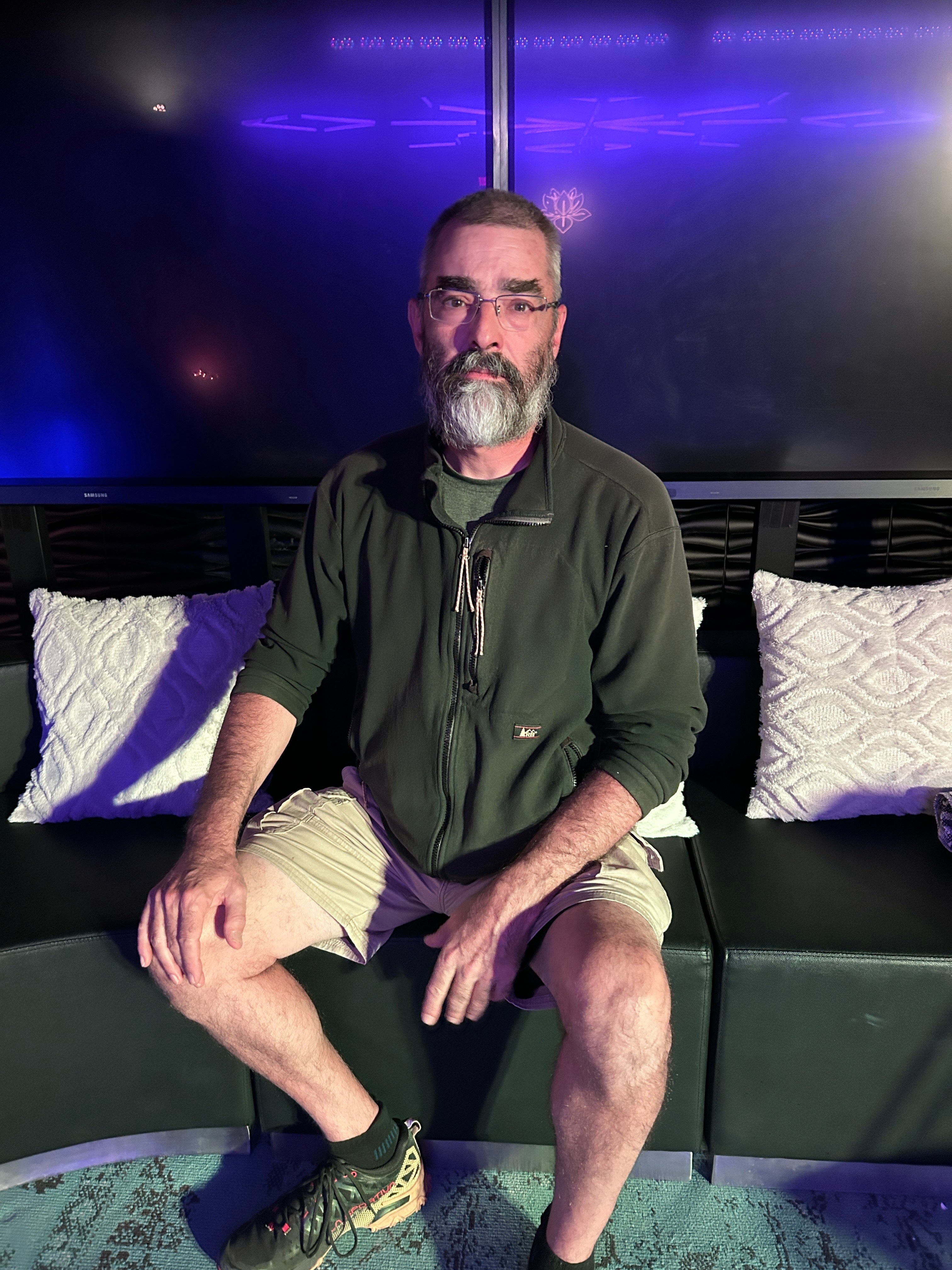 A middle-aged white man with glasses and a beard sitting on a black couch and staring ahead.