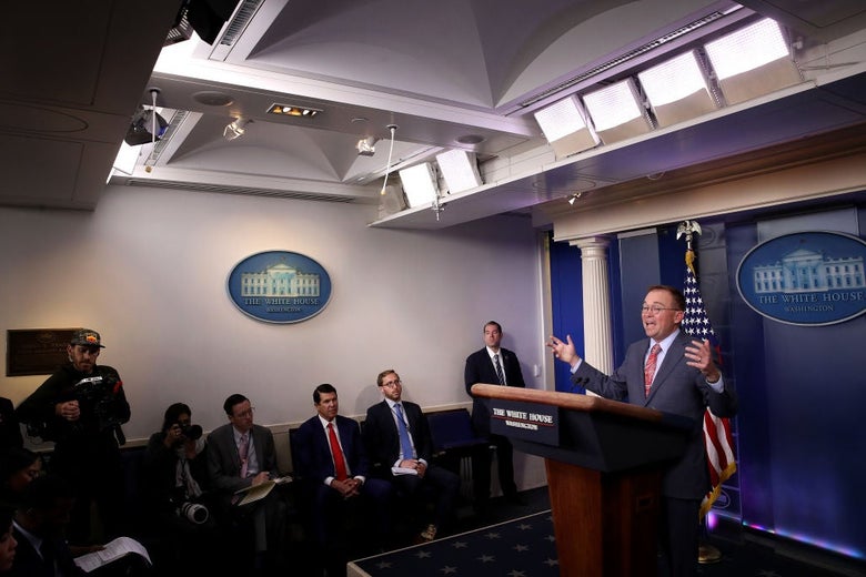 Mulvaney, seen from the side with other individuals seated along a side wall behind him,  gestures while speaking at a White House lectern.