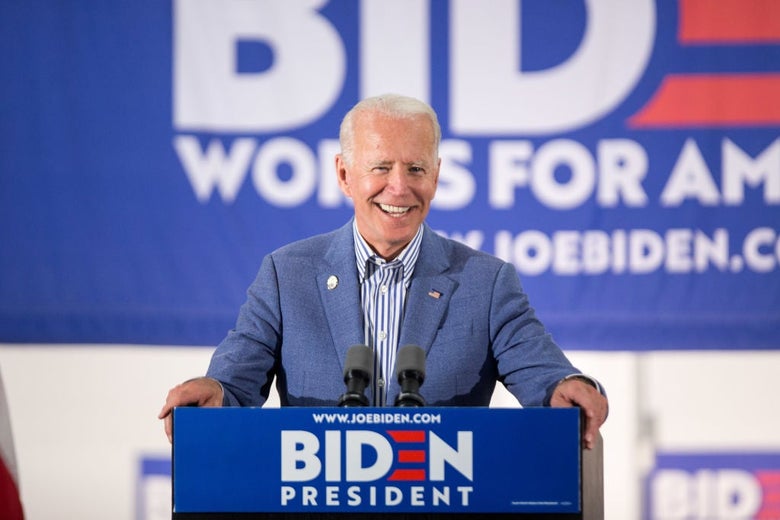 Biden, smiling and wearing a light blue suit with no tie, speaks from a lectern.