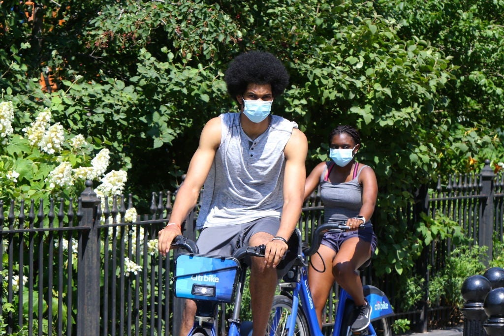 Allen, wearing a sleeveless shirt and a medical mask, rides a blue CitiBike on a path past a garden.