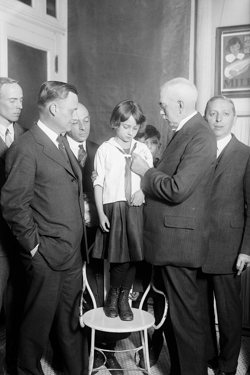 A girl stands on a chair surrounded by men while a man puts a button on her shirt.