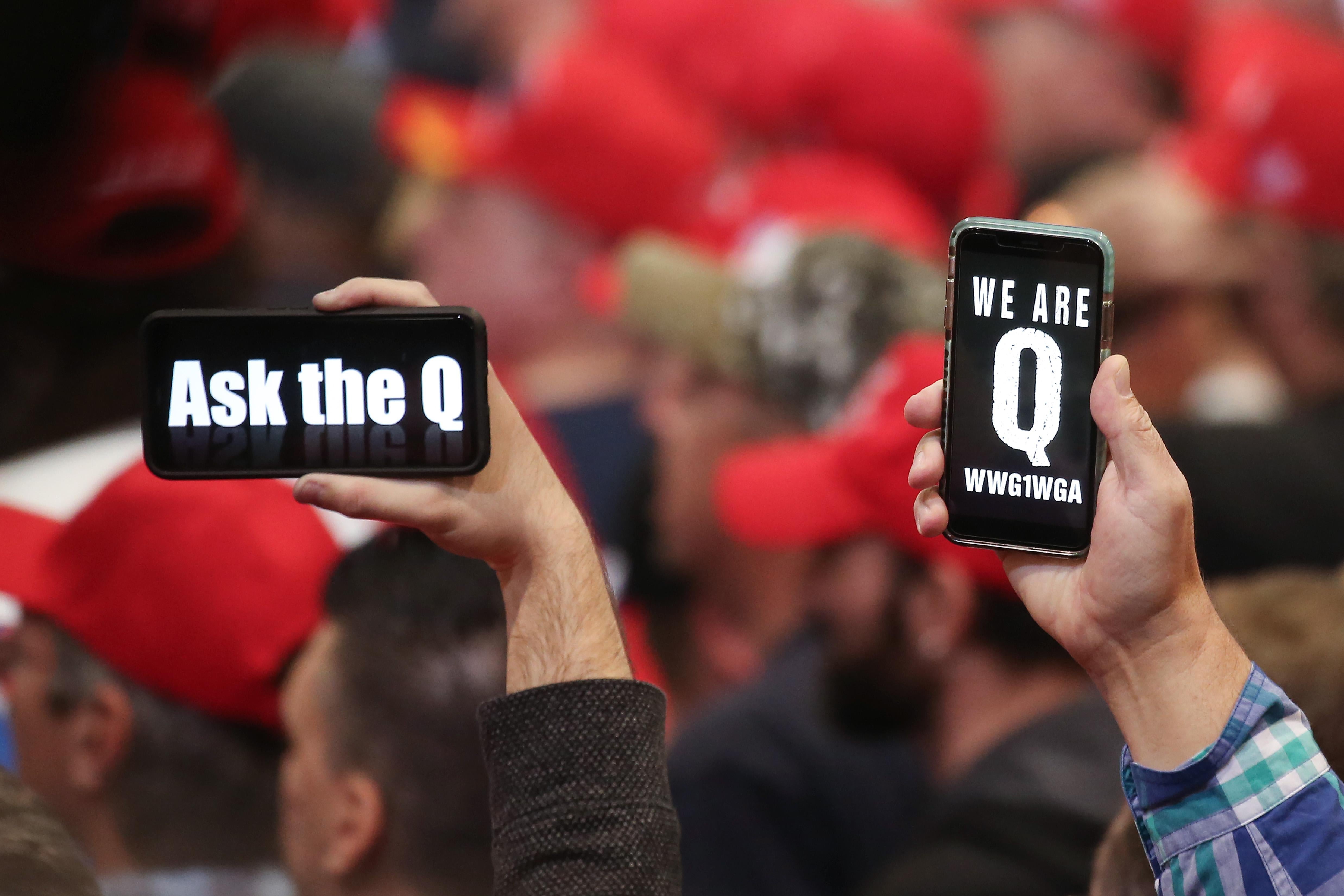 Trump supporters hold up phones displaying QAnon slogans, a sea of red MAGA hats in the background