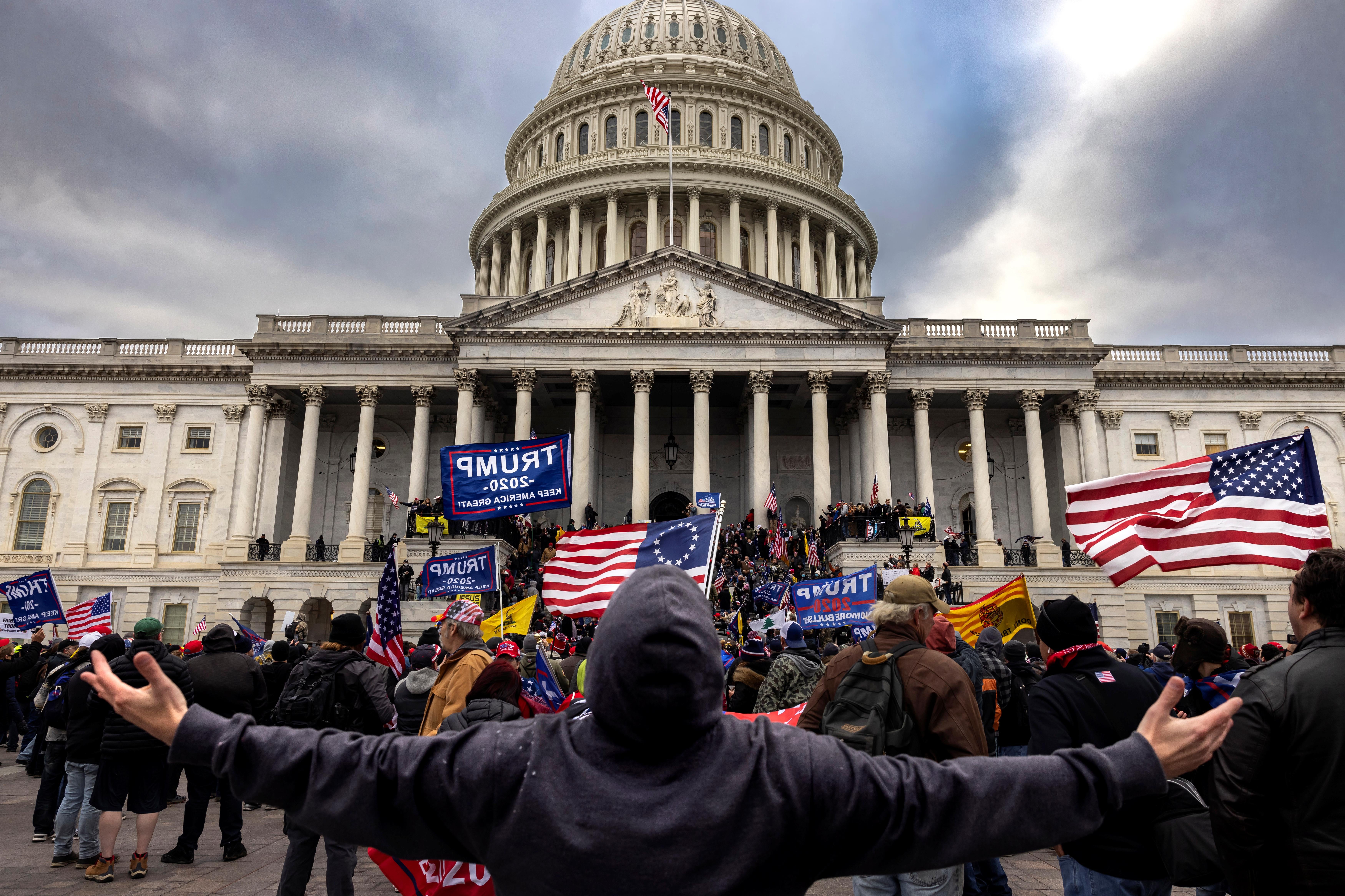 A man in a hoodie stands extending both arms straight out to his sides in front of a crowd of flag-bearing Trump supporters storming the Capitol.