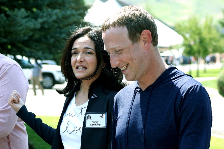 Sheryl Sandberg walks next to Mark Zuckerberg. They are talking, and Sandberg is wearing a name tag. 
