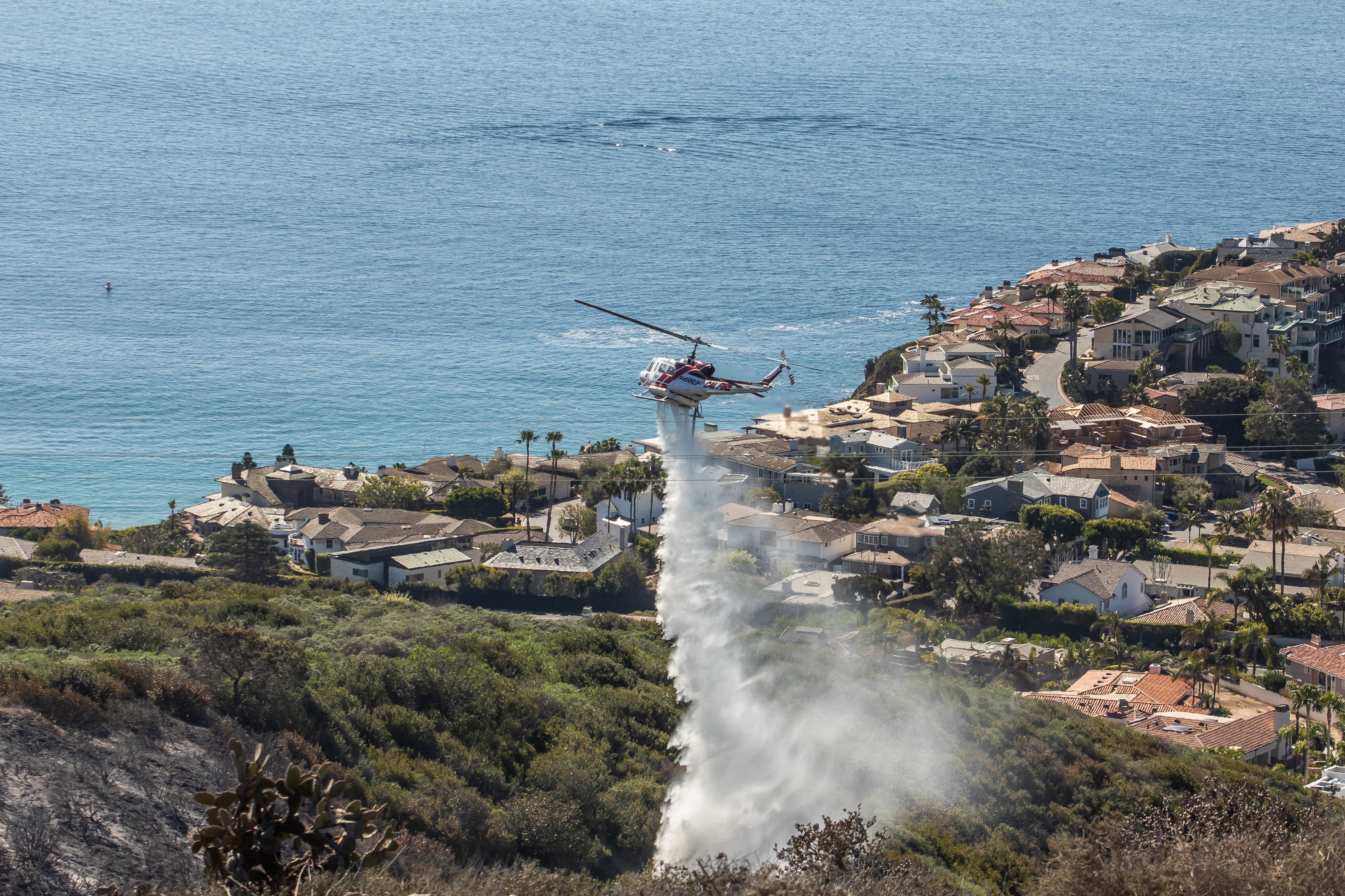 A helicopter drops water on a partially burnt hillside, which overlooks the ocean. Large, evacuated houses are situated nearby the burnt area. 