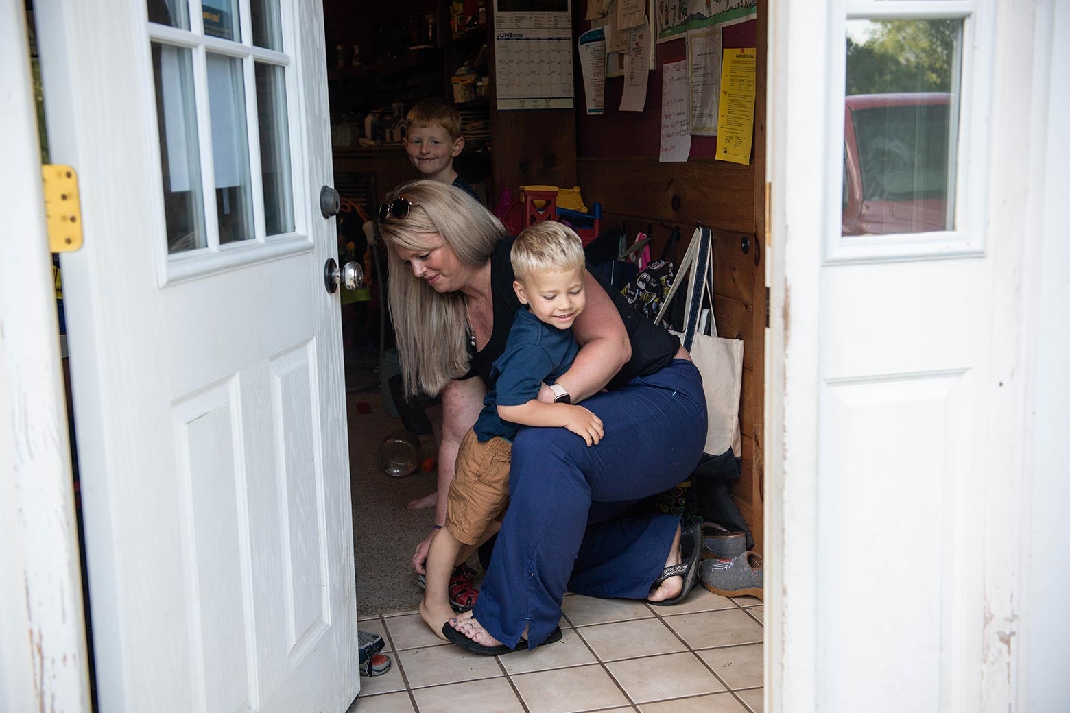 View from outside into Heather's mudroom, where she helps one son take off his shoes as her other son smiles at the camera