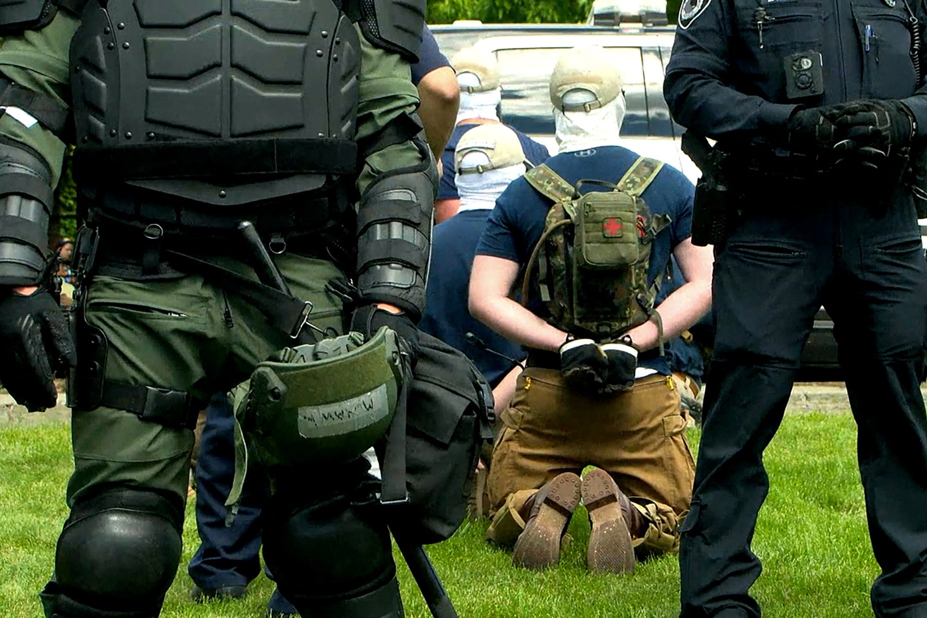 Several men seen from the back wearing baseball caps and white head coverings kneel on the grass in handcuffs surrounded by police in riot gear