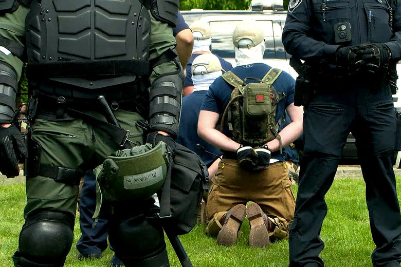 Several men seen from the back wearing baseball caps and white head coverings kneel on the grass in handcuffs surrounded by police in riot gear