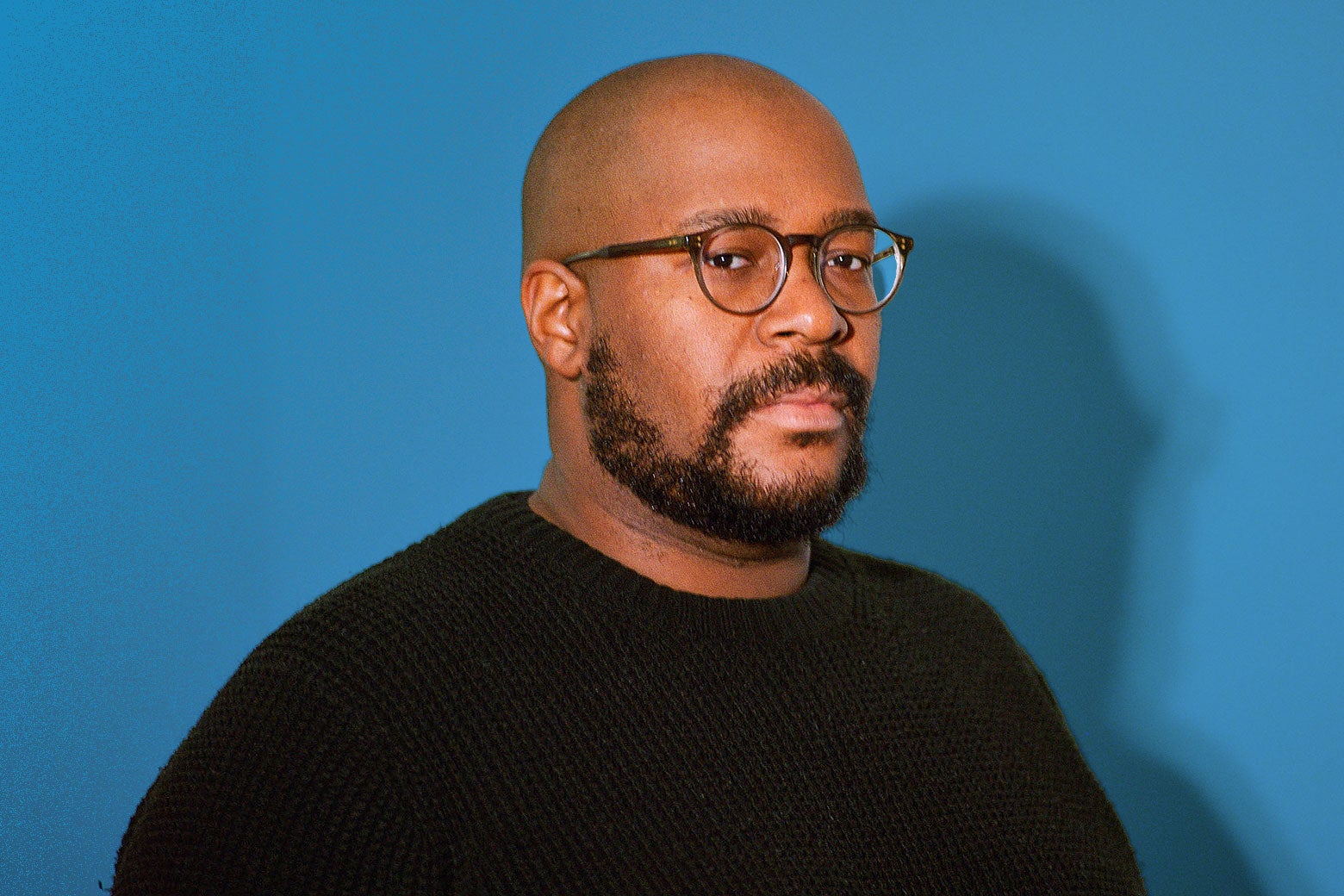 Writer Brandon Taylor, against a blue background, wears framed glasses and a black knit sweater.