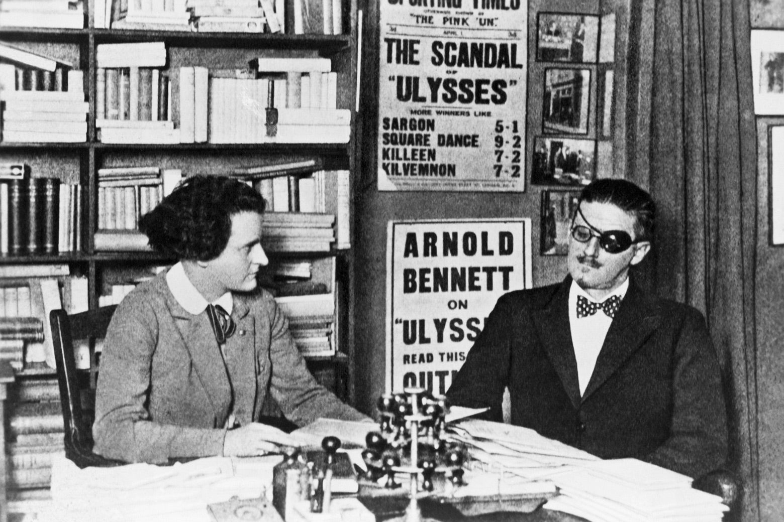 A black-and-white photo of Joyce and Beach sitting at a table in a cluttered office with bookshelves lining the walls. Joyce is wearing an eye patch.