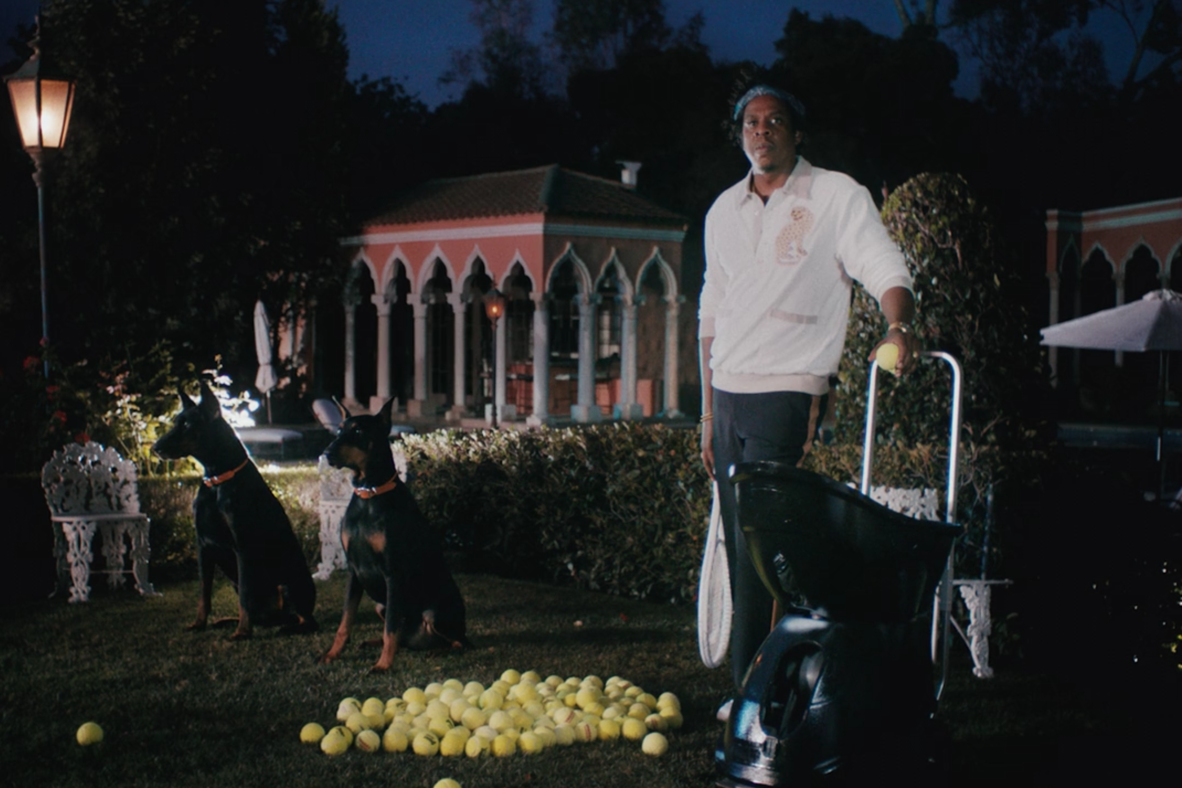 Jay-Z, in tennis gear and holding a racket, stands next to a mound of tennis balls, two dogs, and a ball machine.