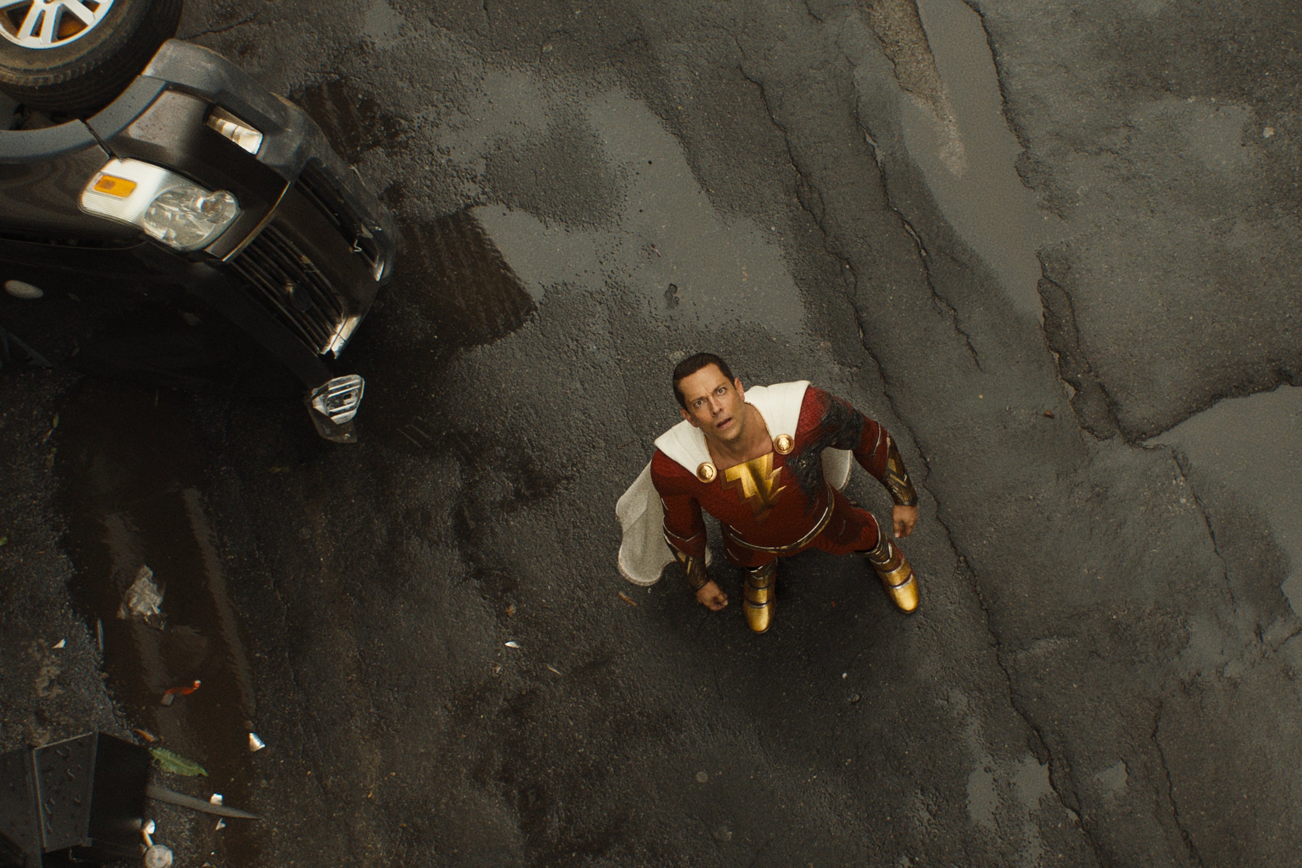 An overhead shot shows the actor standing on the pavement in his red supersuit, next to an overturned car, looking at the sky with concern.