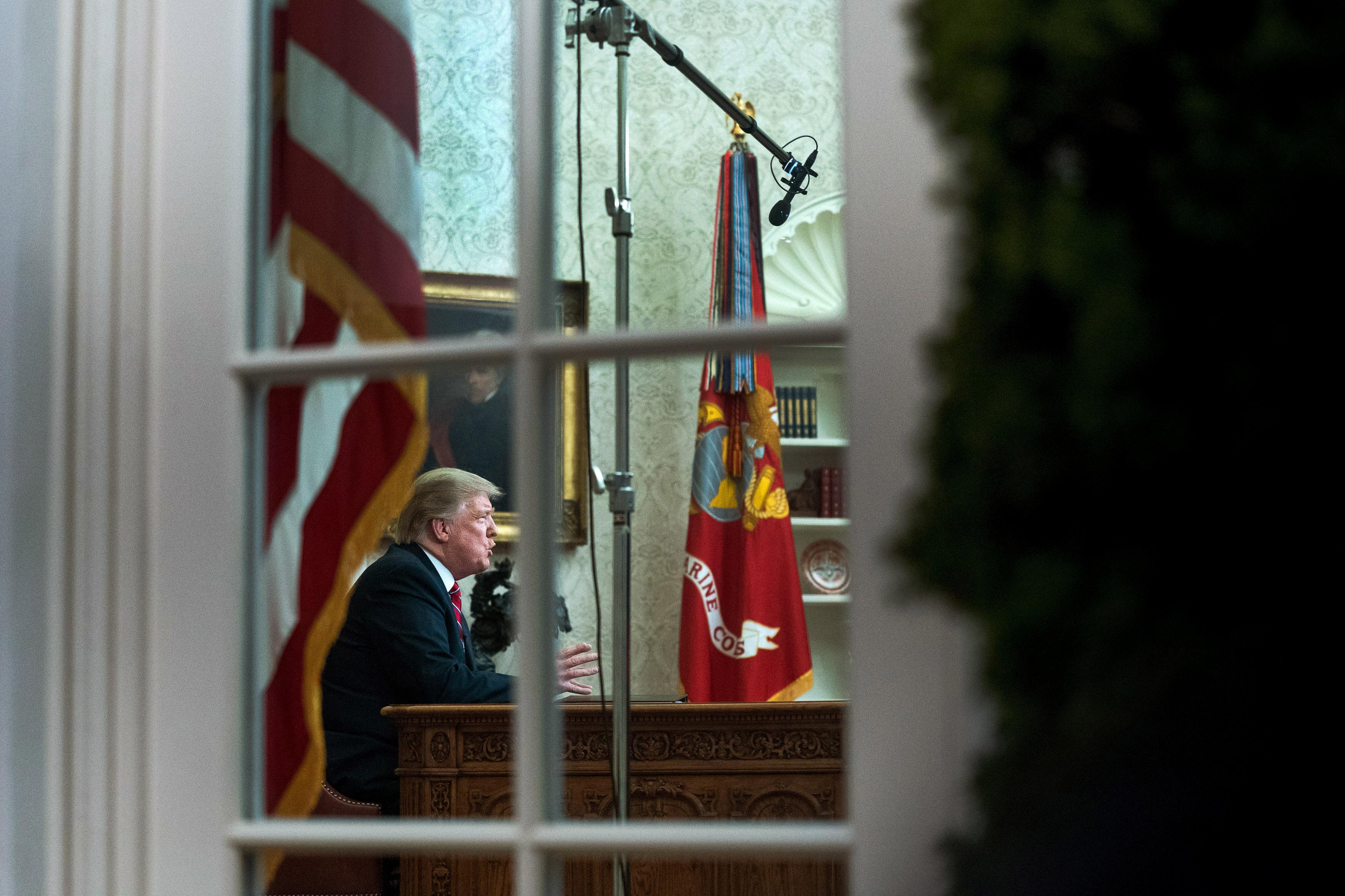 President Donald Trump, seen through the window, speaks to the nation in his first-prime address from the Oval Office of the White House.