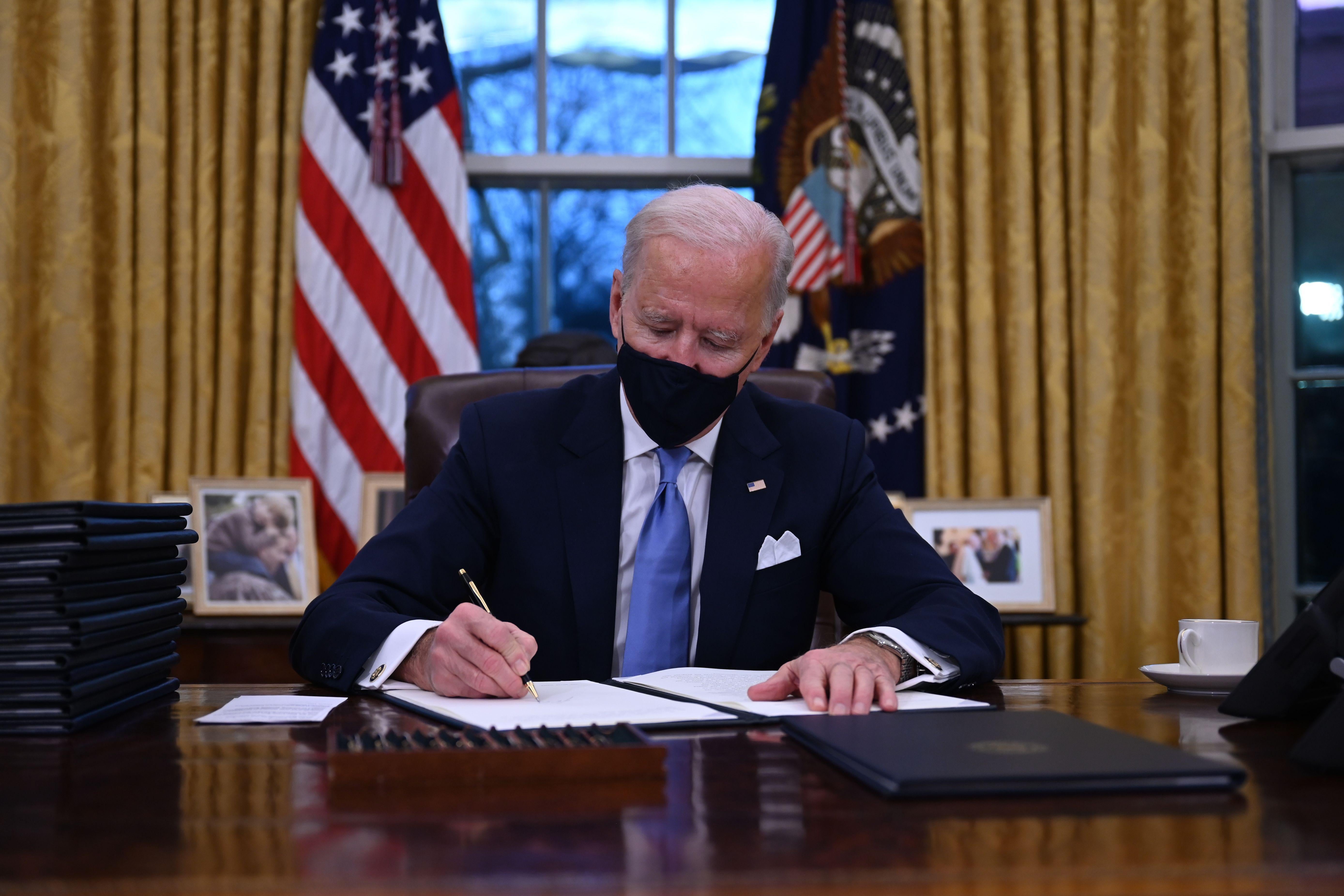 Biden sits in the Oval Office and signs orders.