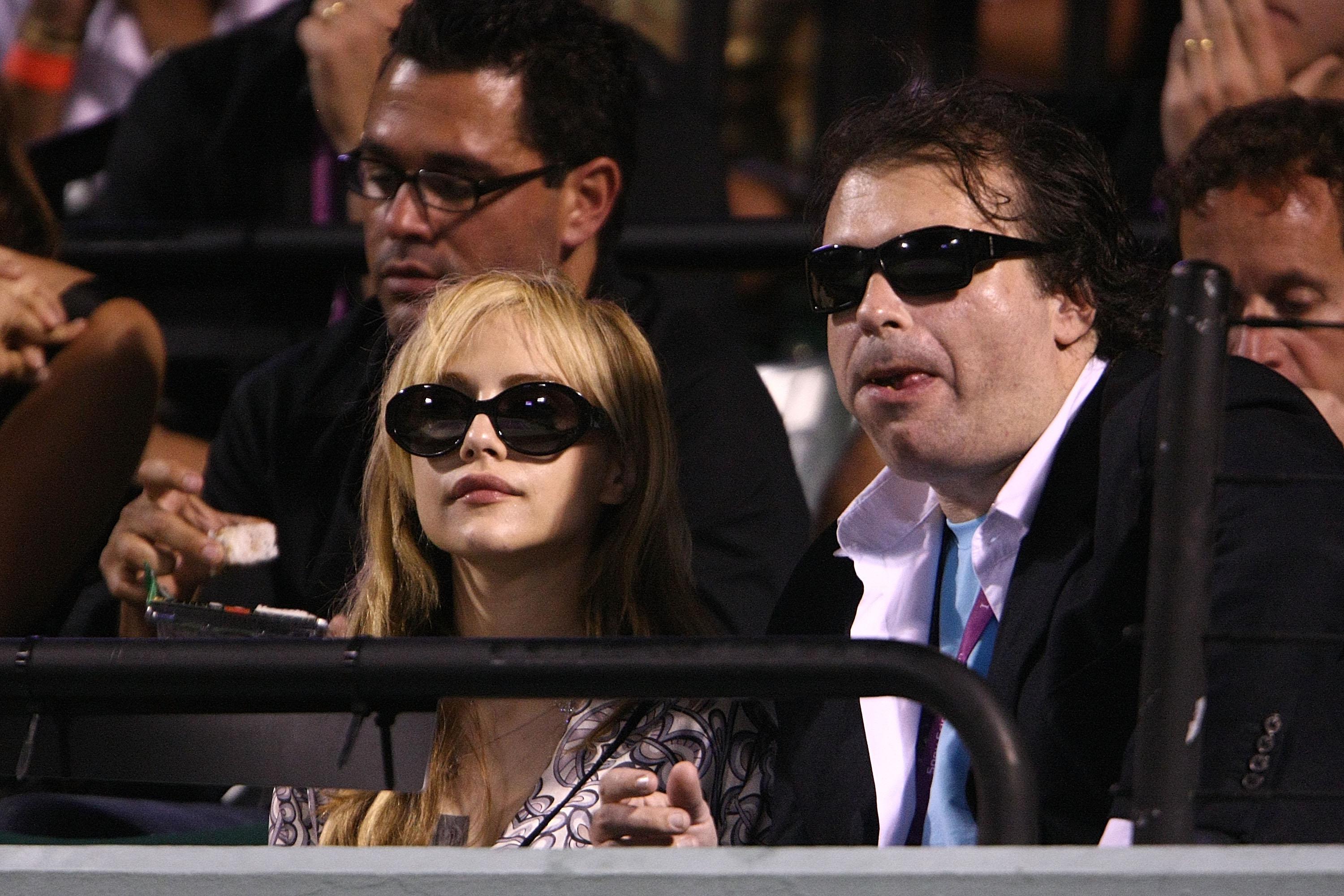 A woman, left, wears black sunglasses and a pink shirt. She is seated with a man, right, in a black jacket, pink button down shirt, and blue t-shirt; he also wears sunglasses. They are looking out onto a tennis court off-camera.