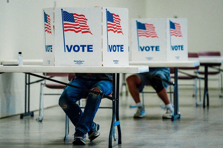 Two people sit behind privacy barriers on spaced-out desks, filling out their ballots