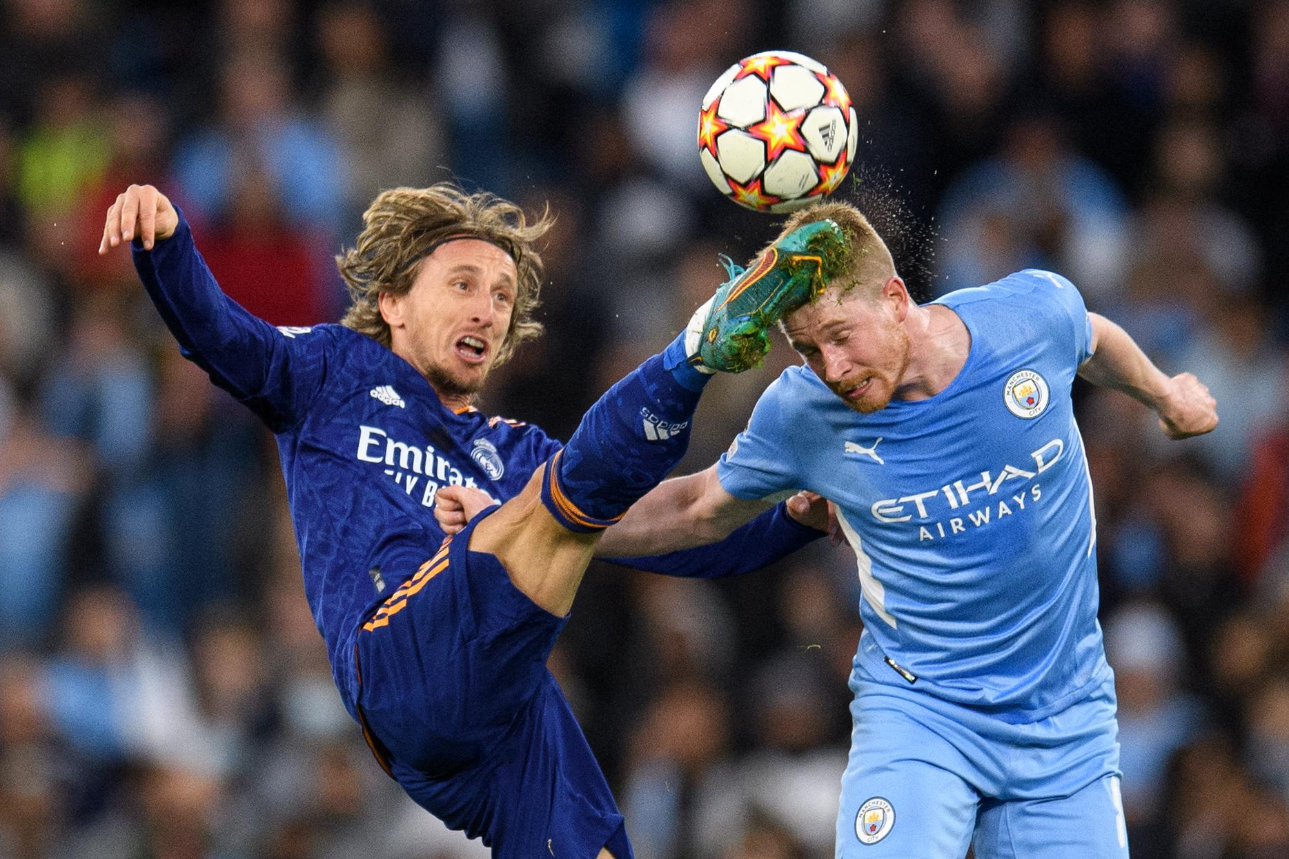 De Bruyne heads the ball with his eyes closed with a fist at Modric's chest as Modric looks up at and kicks toward the ball but hits De Bruyne in the head with his foot