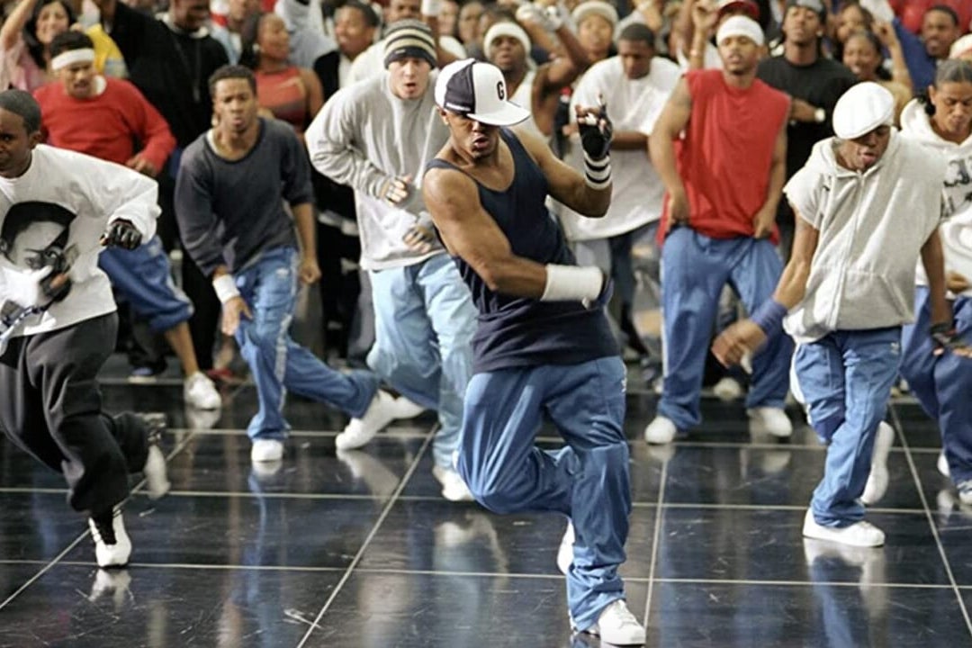 A crew of break dancers are mid-battle in front of a crowd on a sheen floor. 