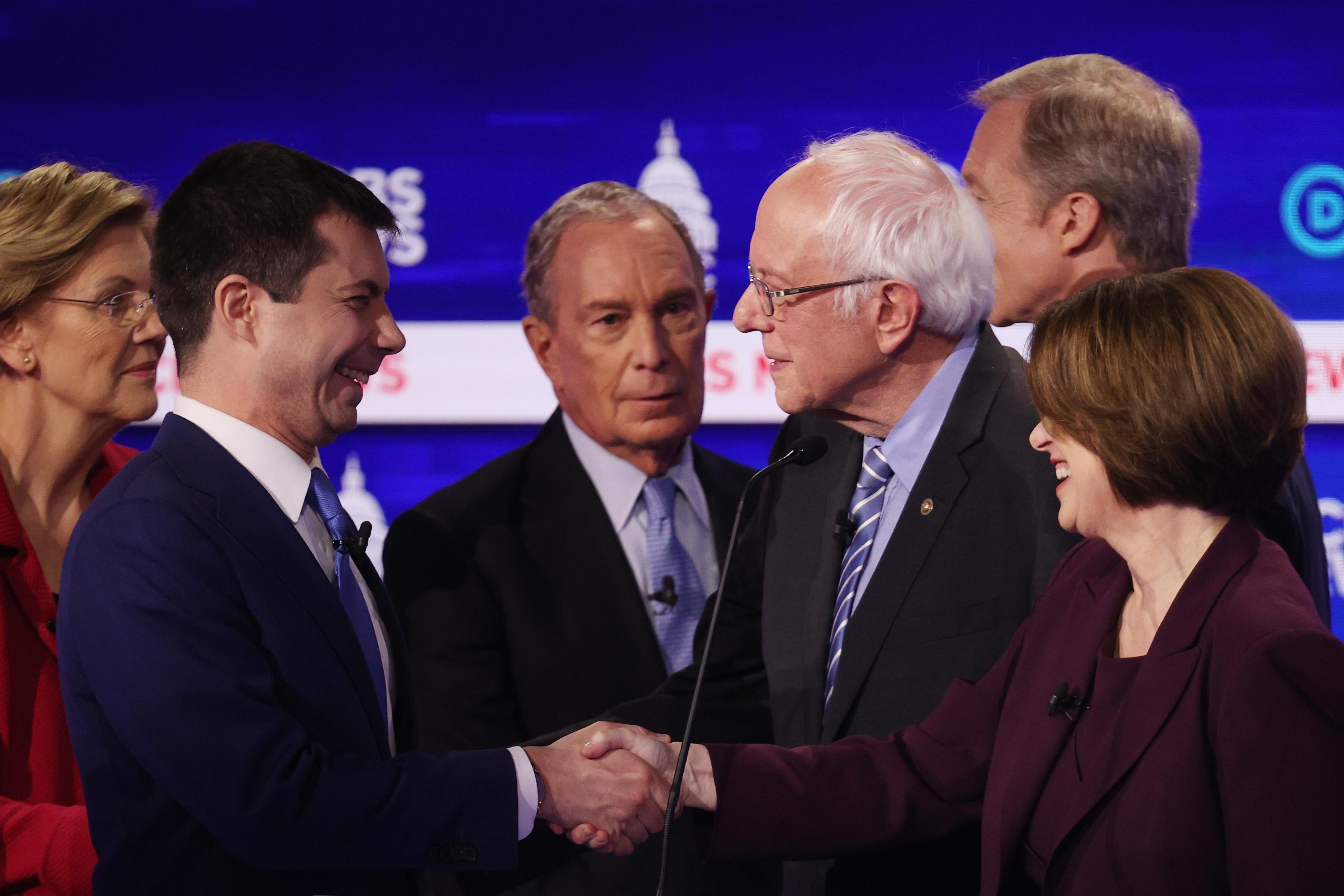 The candidates greet each other on the debate stage. Buttigieg and Klobuchar shake hands, both smiling.