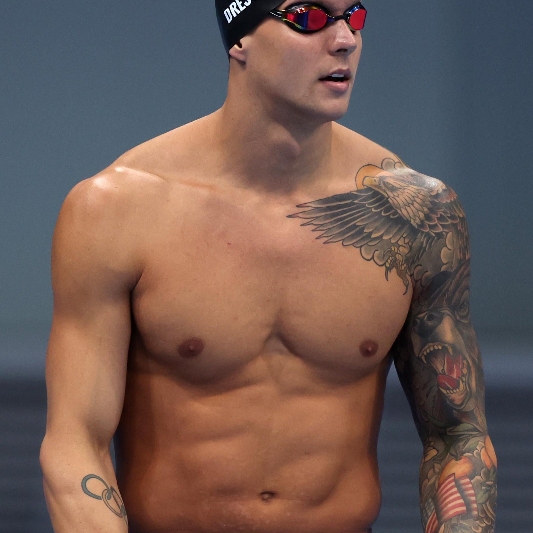 Dressel in goggles and swim cap, his left arm covered in the eagle, bear, oranges, and flag tattoo described below, a small Olympics rings tattoo visible as well on his right forearm