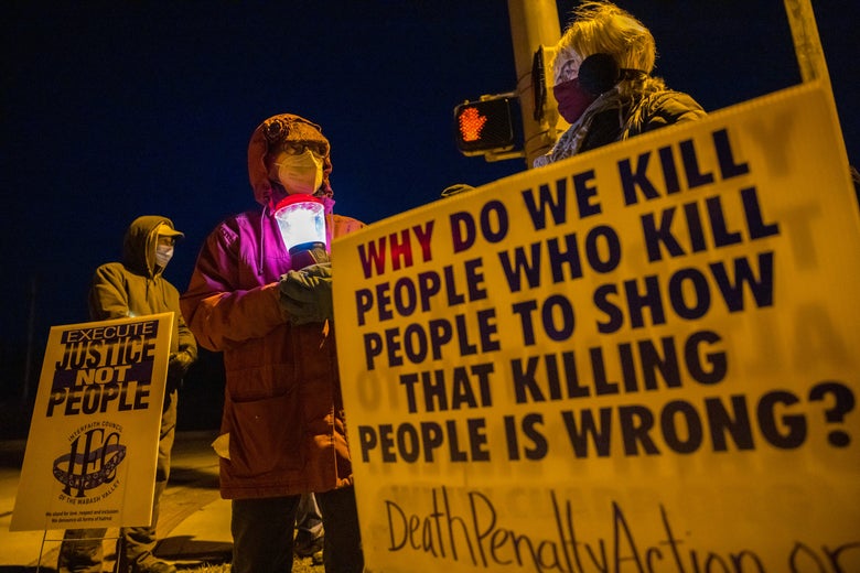 People in jackets hold signs that say "Execute Justice Not People" and "Why do we kill people who kill people to show that killing people is wrong?" outside at night