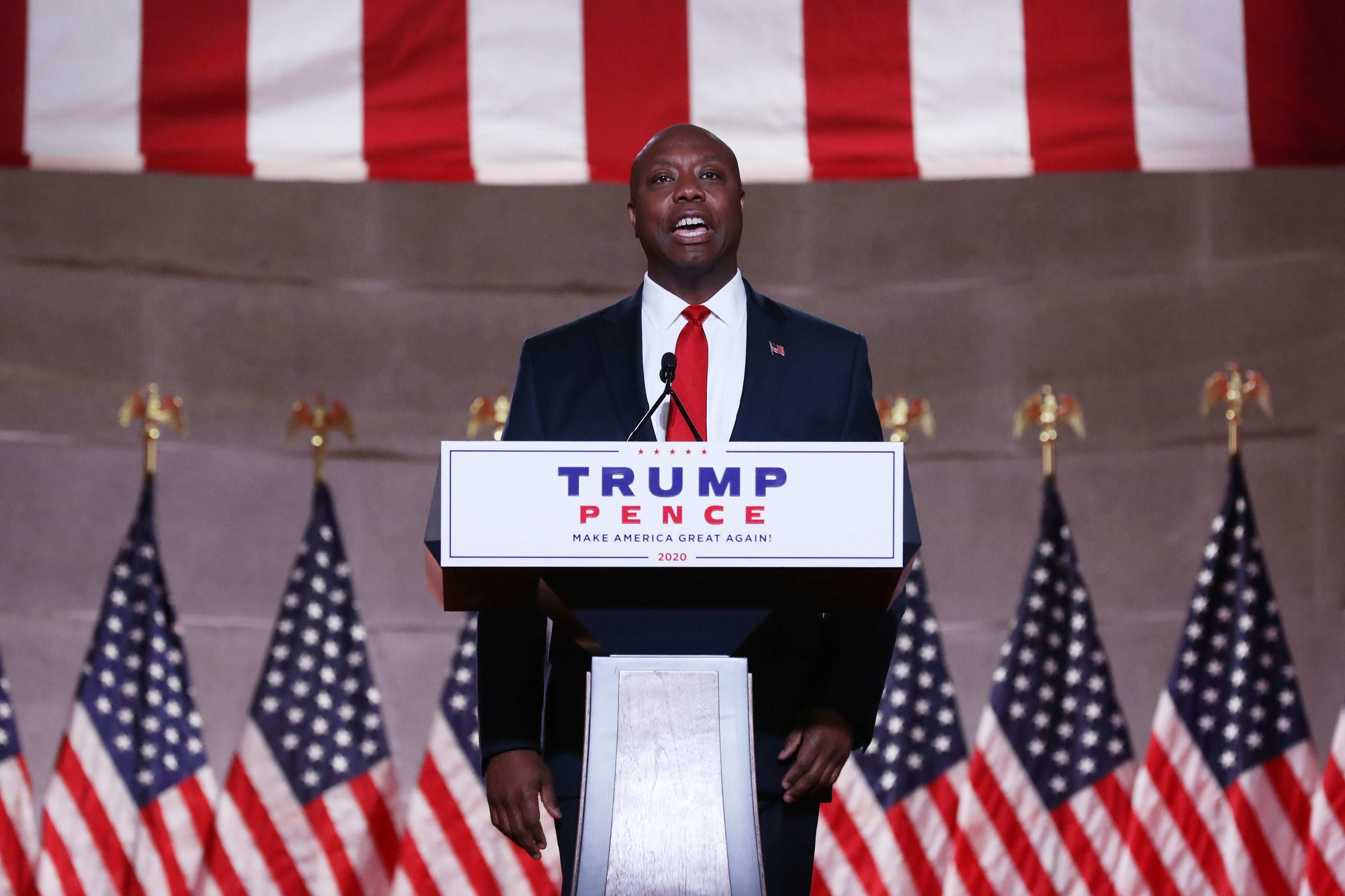 Tim Scott speaks at a podium with a "Trump Pence" sign on it. Behind him are American flags.