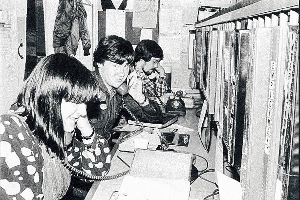 A woman and two men sit at a long desk, with telephones to their ears.