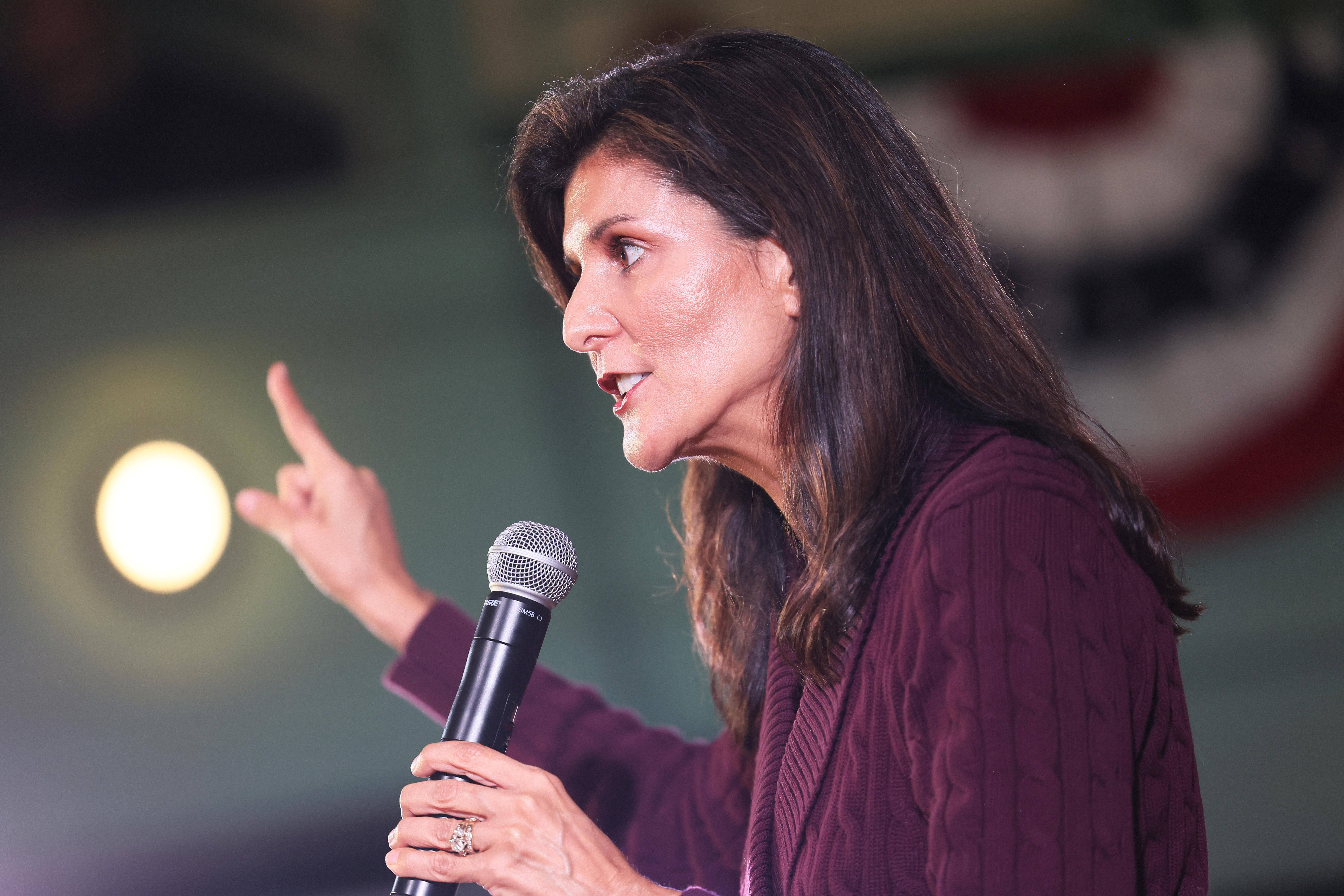 A close-up shot of Nikki Haley, wearing a purple sweater and holding a microphone in one hand.