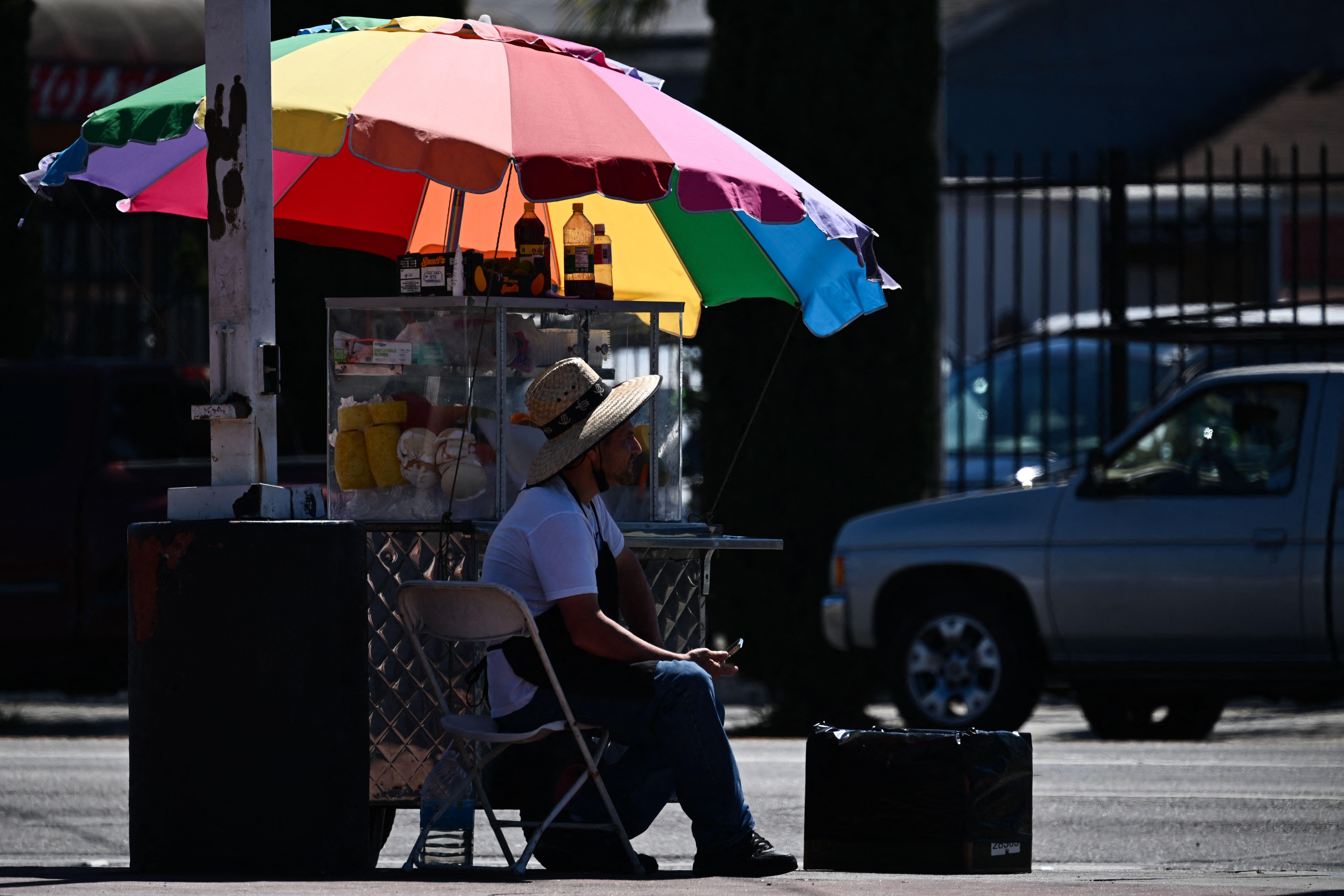 A person in a straw hat sits in the shade under a rainbow-colored umbrella.