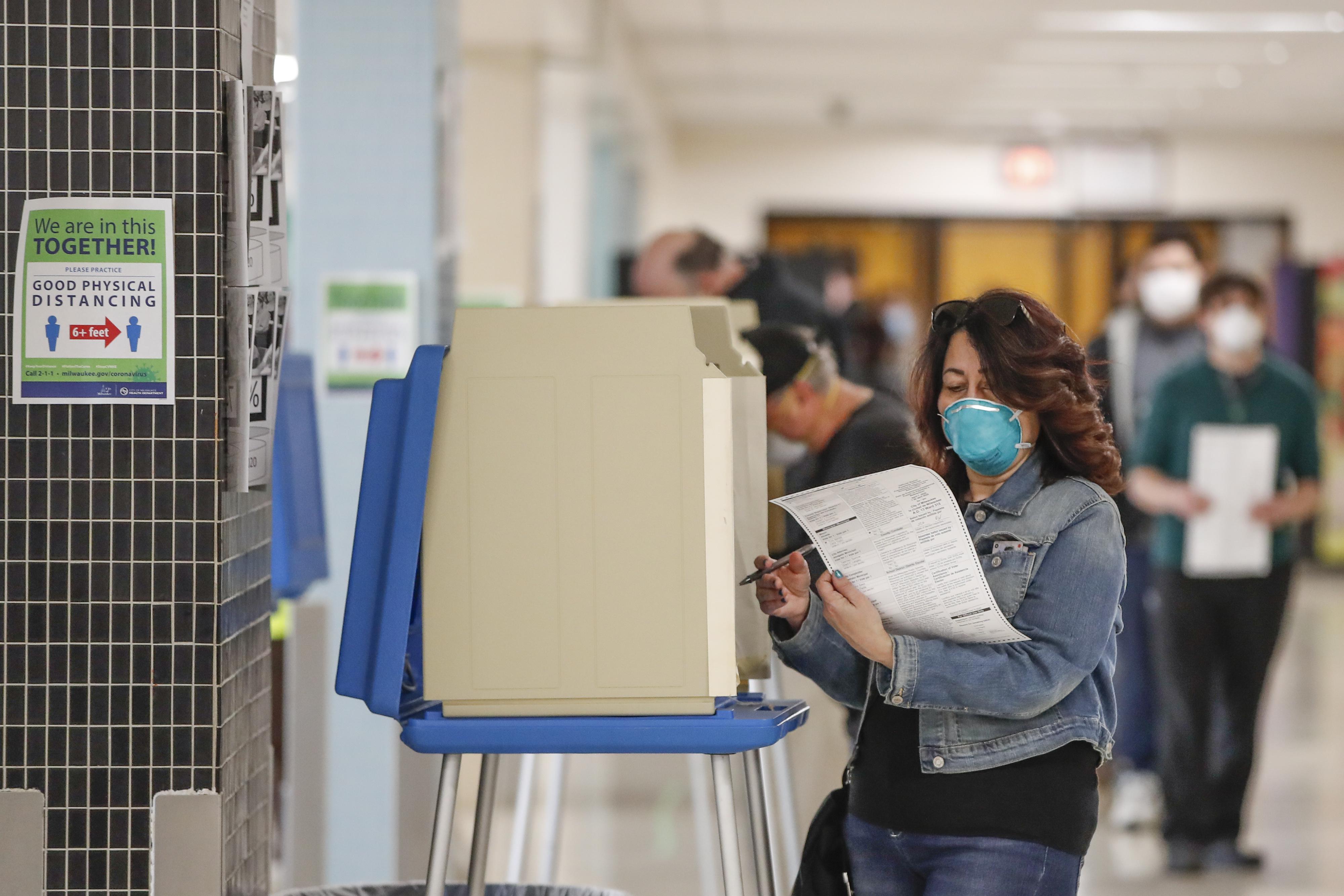 A woman wearing a mask looks at her ballot, with other voters wearing masks in the background