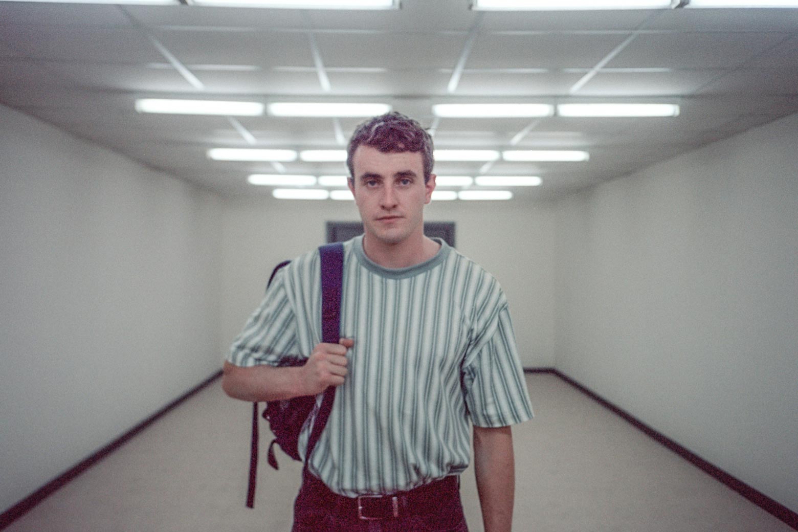 The handsome actor walks down a sterile looking white hallway, fluorescent lighting overhead, a somber expression on his face, one strap of a backpack over his tucked-in '90s-looking vertical stripe t-shirt