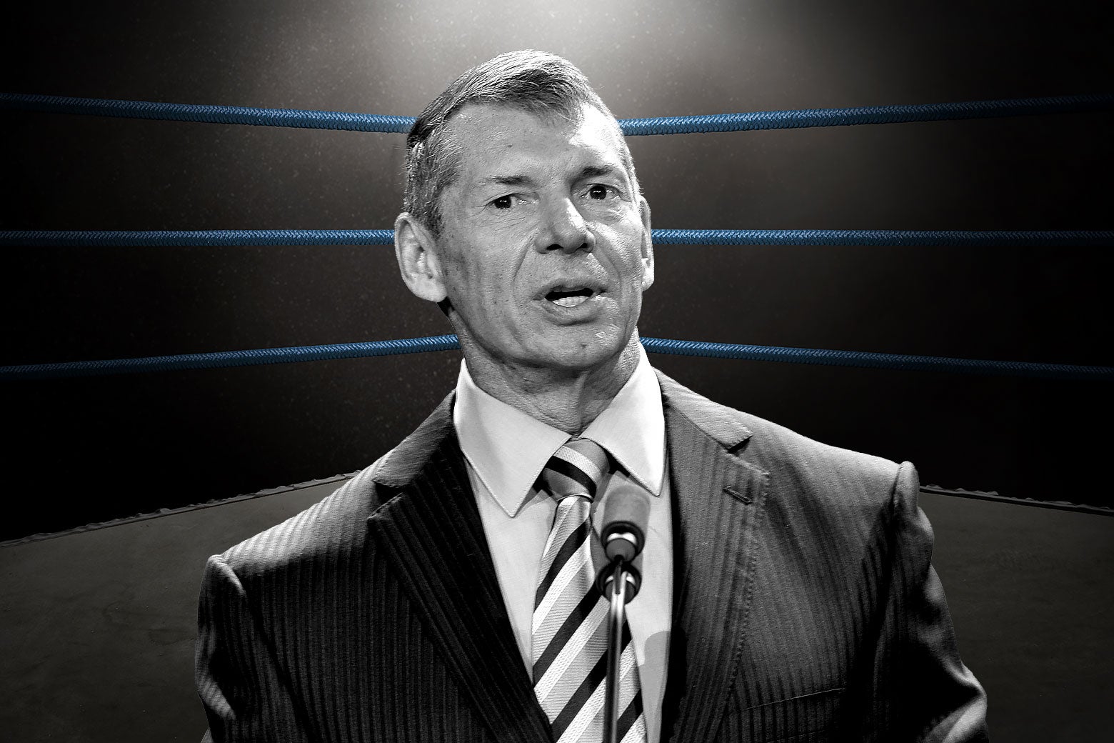 Black-and-white image of Vince McMahon wearing a pinstripe suit and standing behind a microphone in the center of a wrestling ring.
