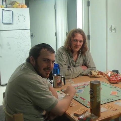 A young man in a kippah and a young red-headed, long-haired man (Black) look at the camera. They are seated around a kitchen table in a white, sparse room, with pringles, a monopoly board, and other snacks.