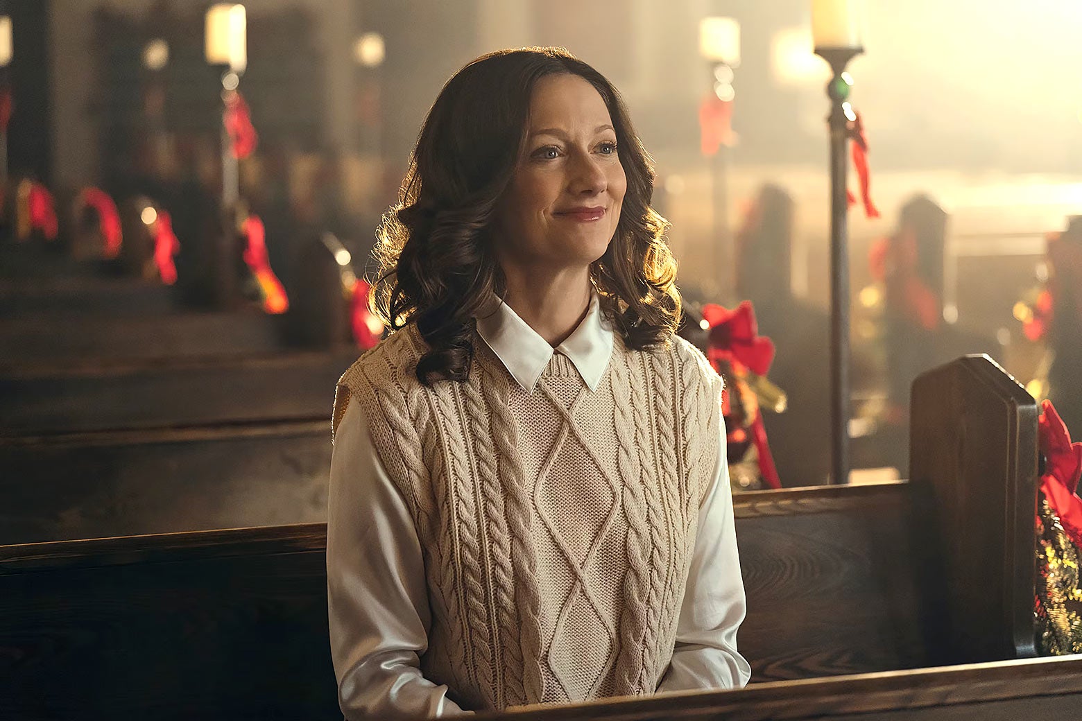 She sits in the front pew of an empty chapel, red ribbons all around her, her hair in curls, wearing a sweater vest over a white blouse. She gazes ahead, glowing.