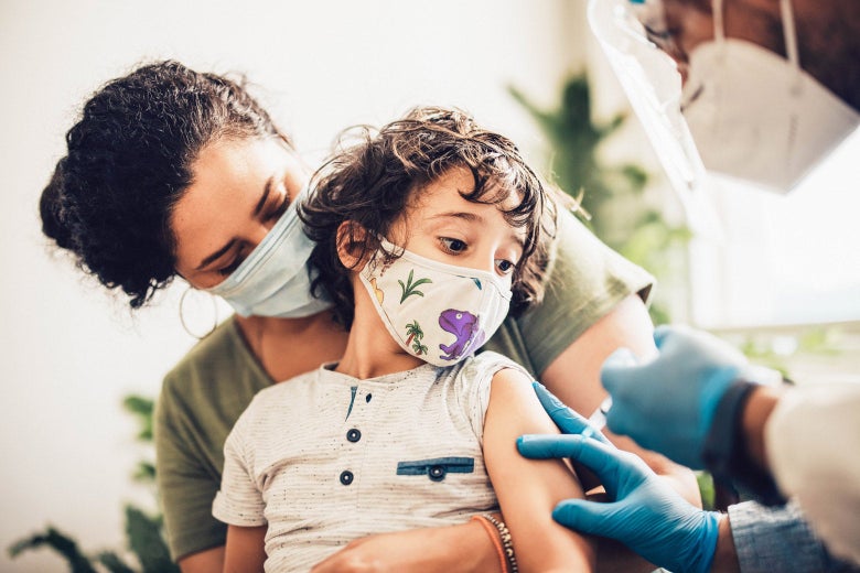 A child in a mask sits on the lap of a woman in a mask as the child gets a vaccine.