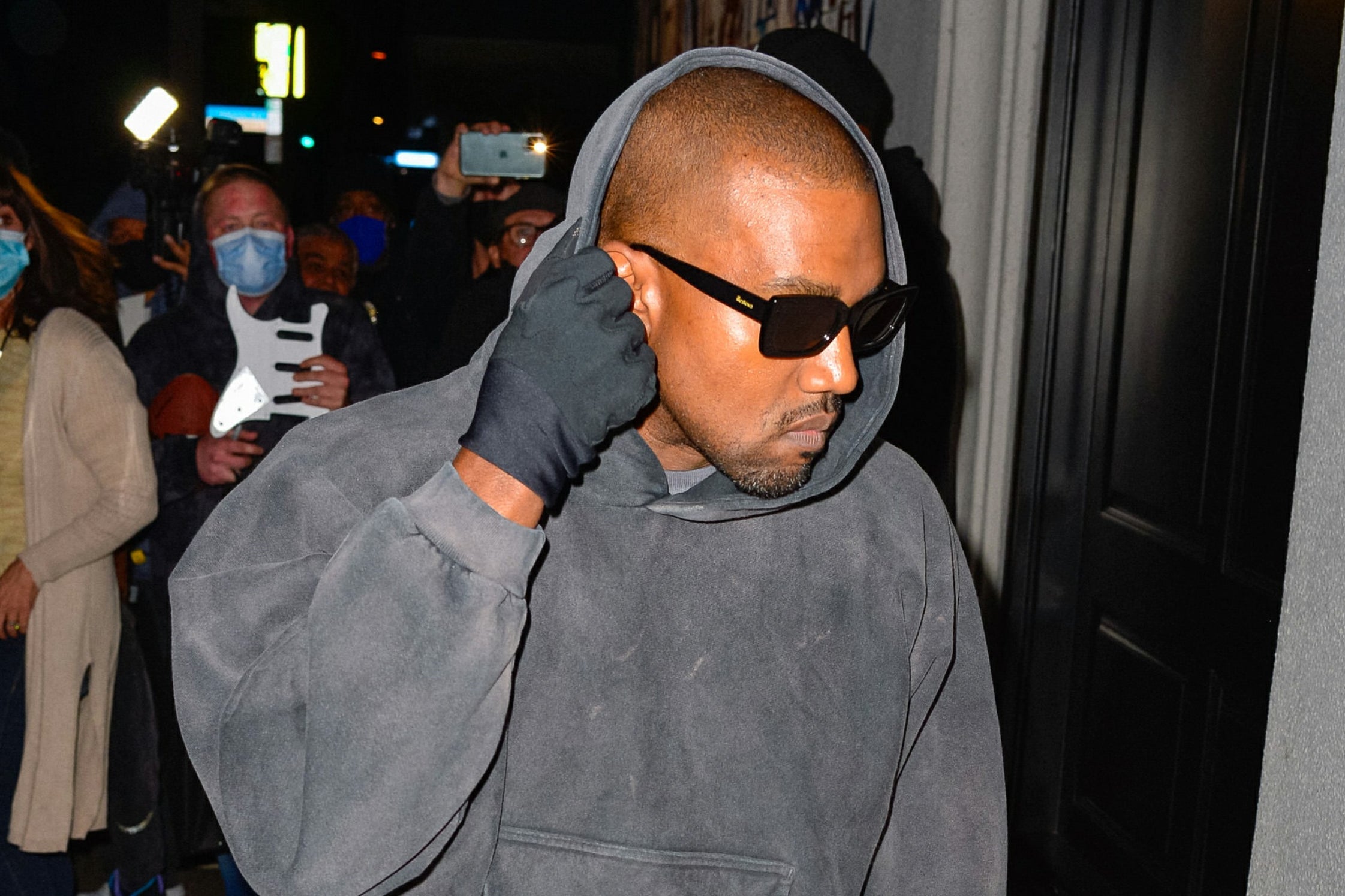 A photo of Kanye West, walking into a restaurant at night, wearing sunglasses, gloves and a hooded sweatshirt.