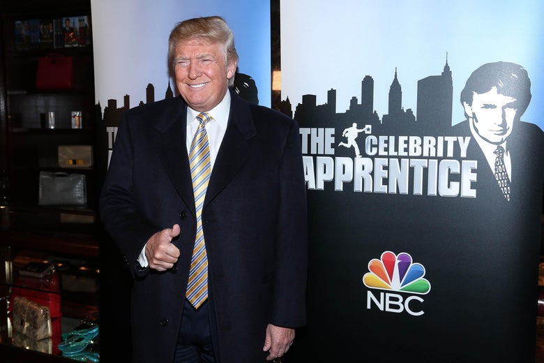 Donald Trump gives a thumbs-up and smiles while standing in front of a Celebrity Apprentice poster.