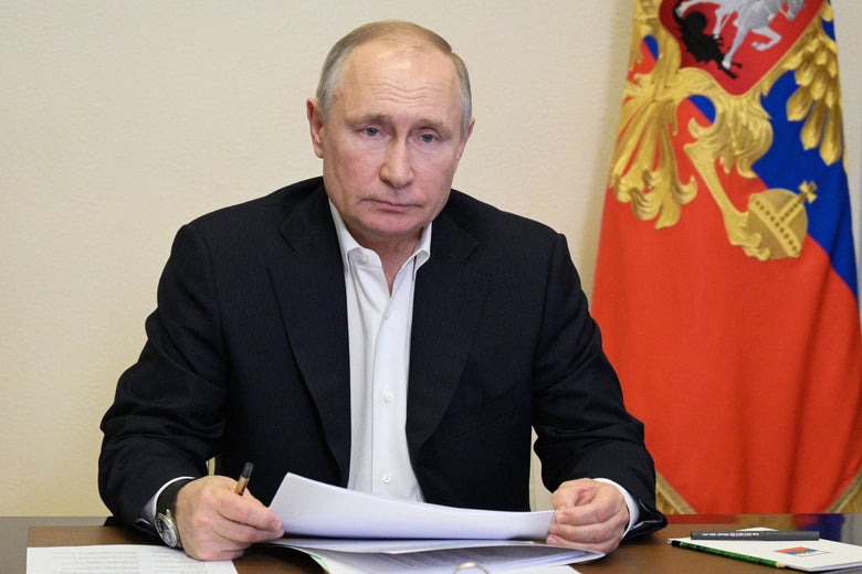 Putin sits at a desk and holds some documents. A flag is seen at his right.