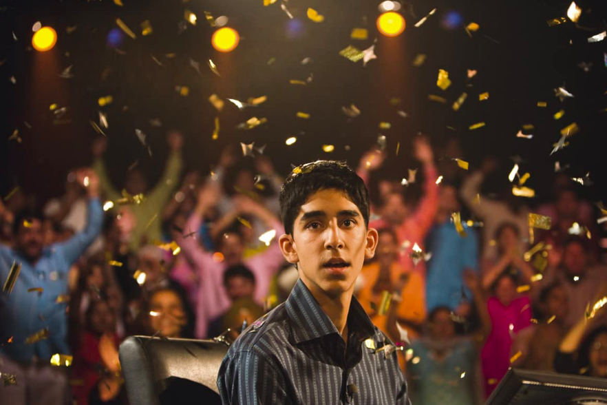 Dev Patel looks directly into the camera, confetti raining down as a crowd cheers behind him.