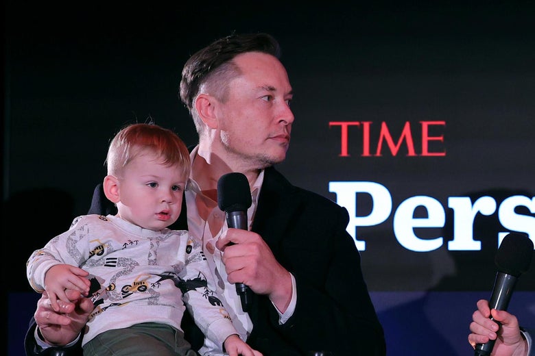 A seated Elon Musk holds a microphone in his hand and a child in his lap.