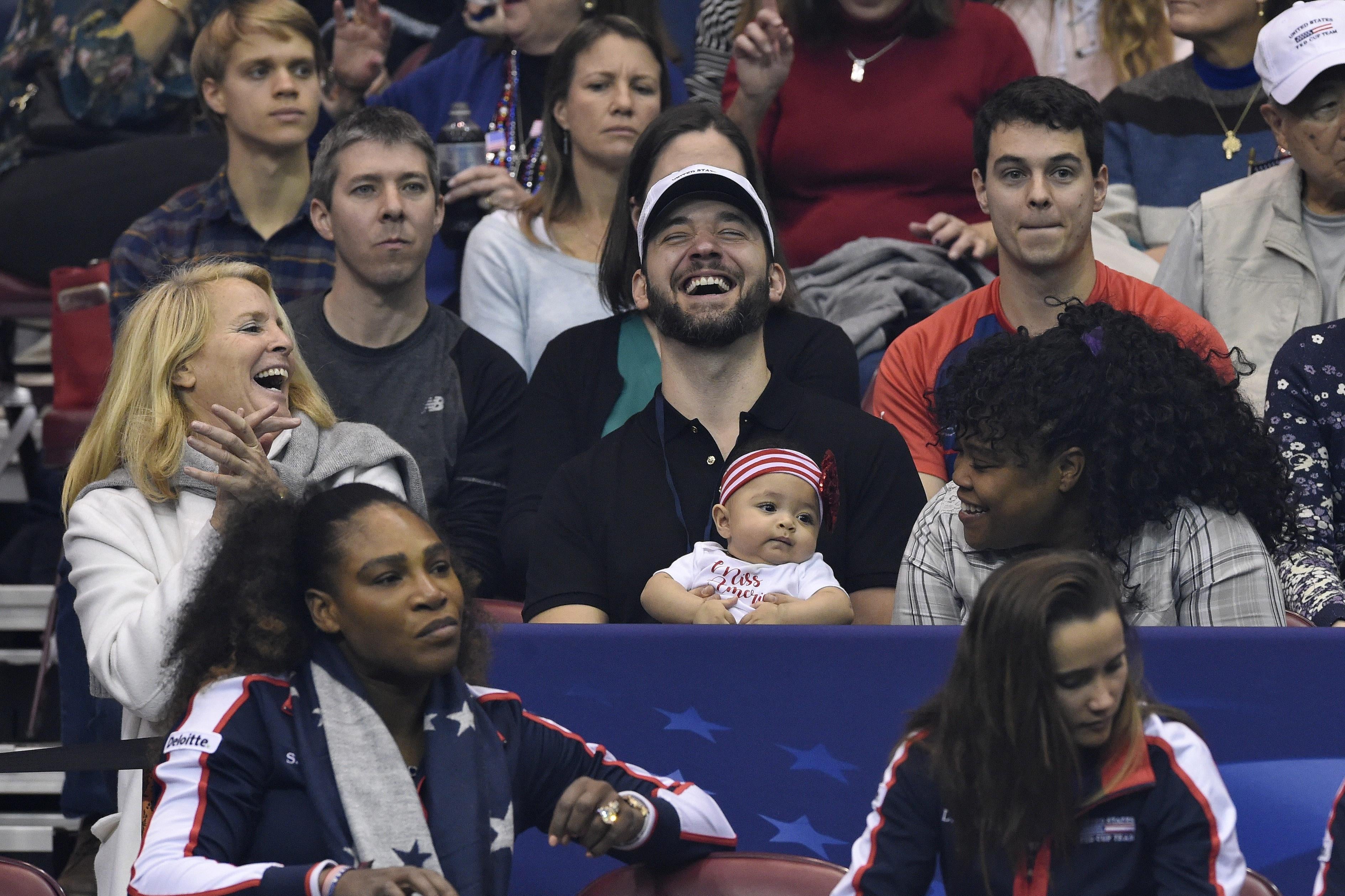 Serena Williams and her husband and child watch a tennis match.