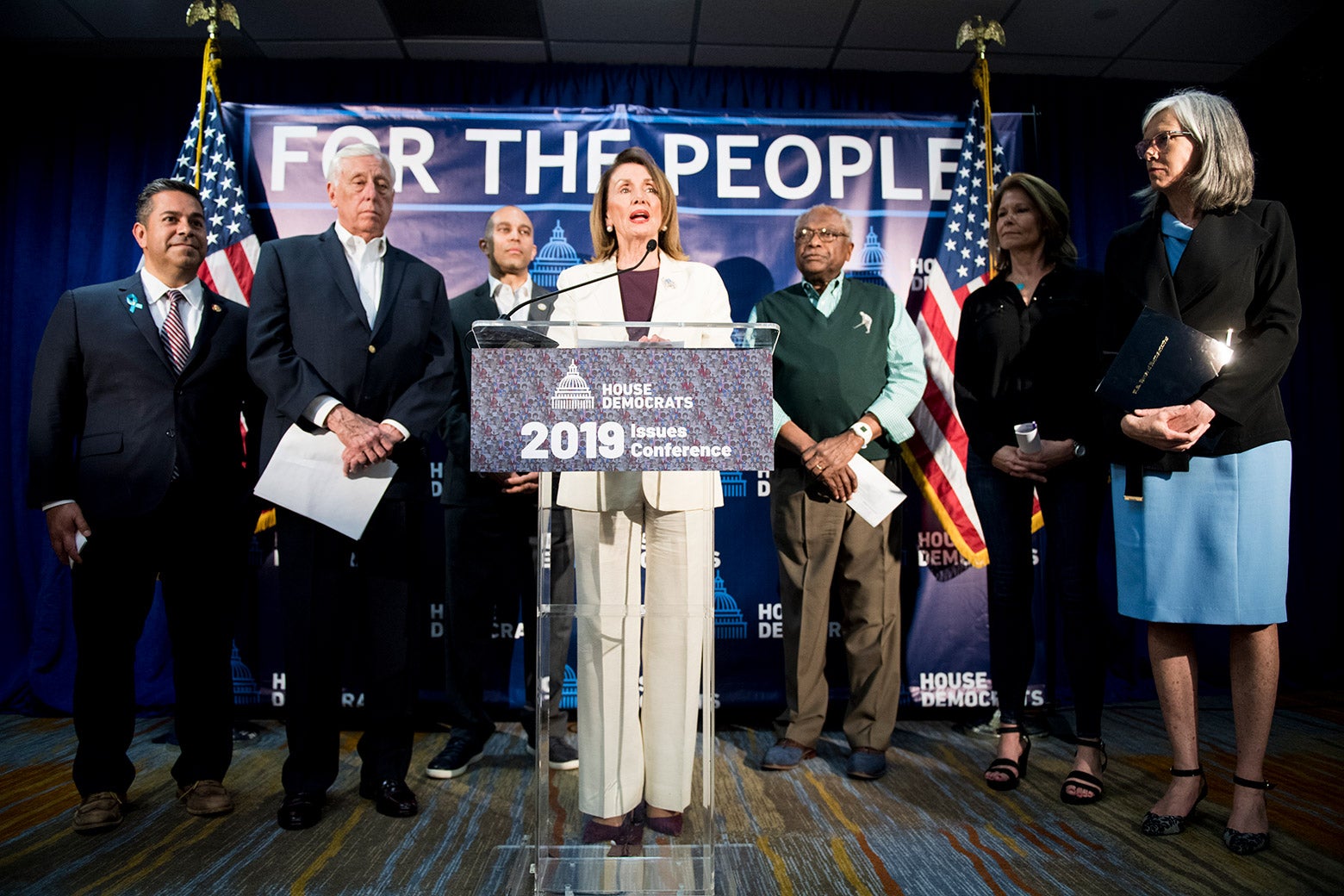 Speaker of the House Nancy Pelosi speaking at a podium, flanked from left by Assistant Democratic Leader Ben Ray Luján, House Majority Leader Steny Hoyer, Democratic Caucus Chair Hakeem Jeffries, House Majority Whip Jim Clyburn, Democratic Congressional Campaign Committee Chair Cheri Bustos, and Democratic Caucus Vice Chair Katherine Clark.