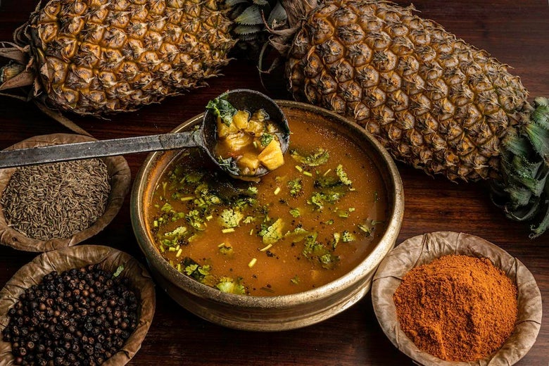 A pot of rasam surrounded by pineapples and bowls of different spices on a wooden surface