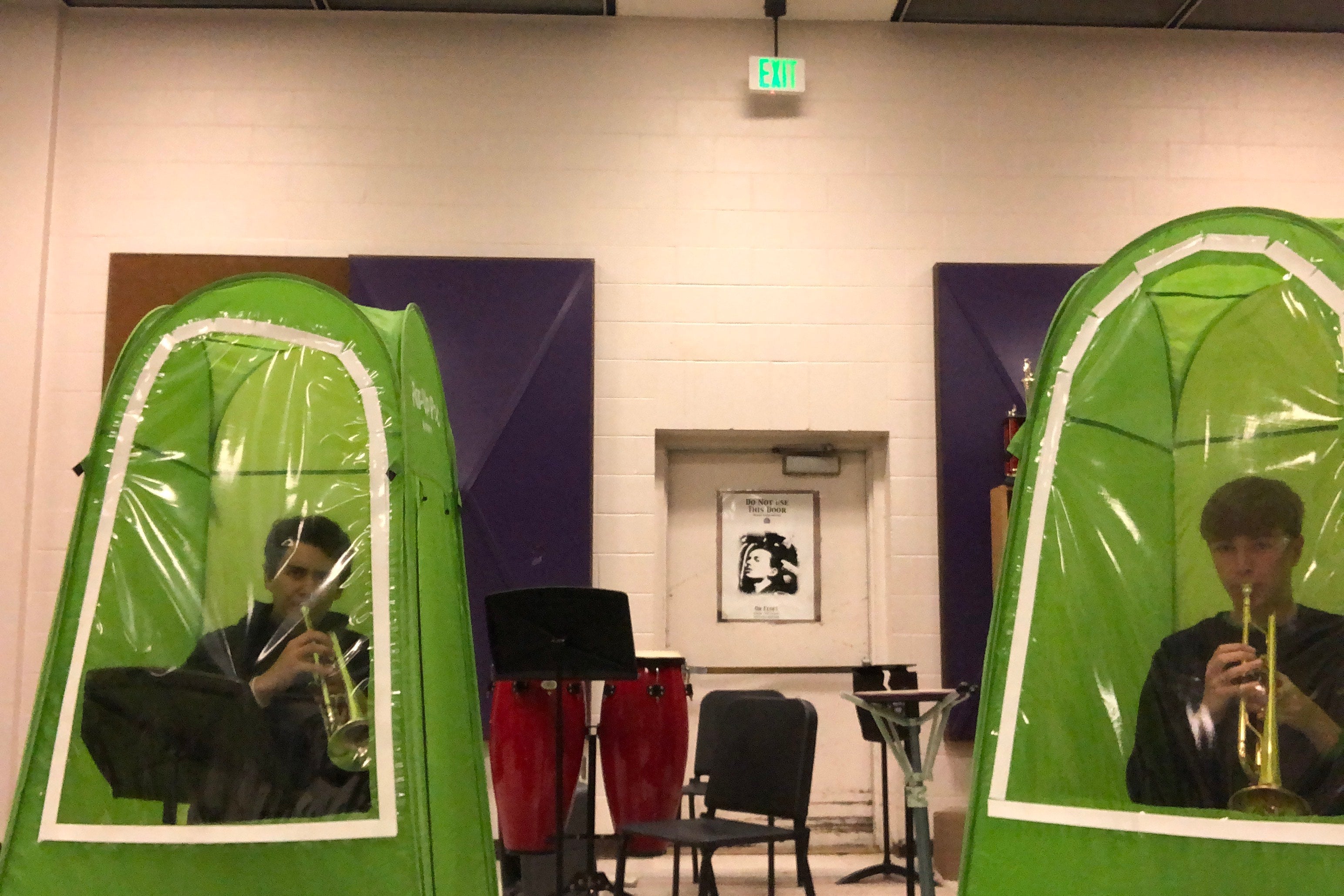 Two trumpet players practice in separate green tents inside a school band room.