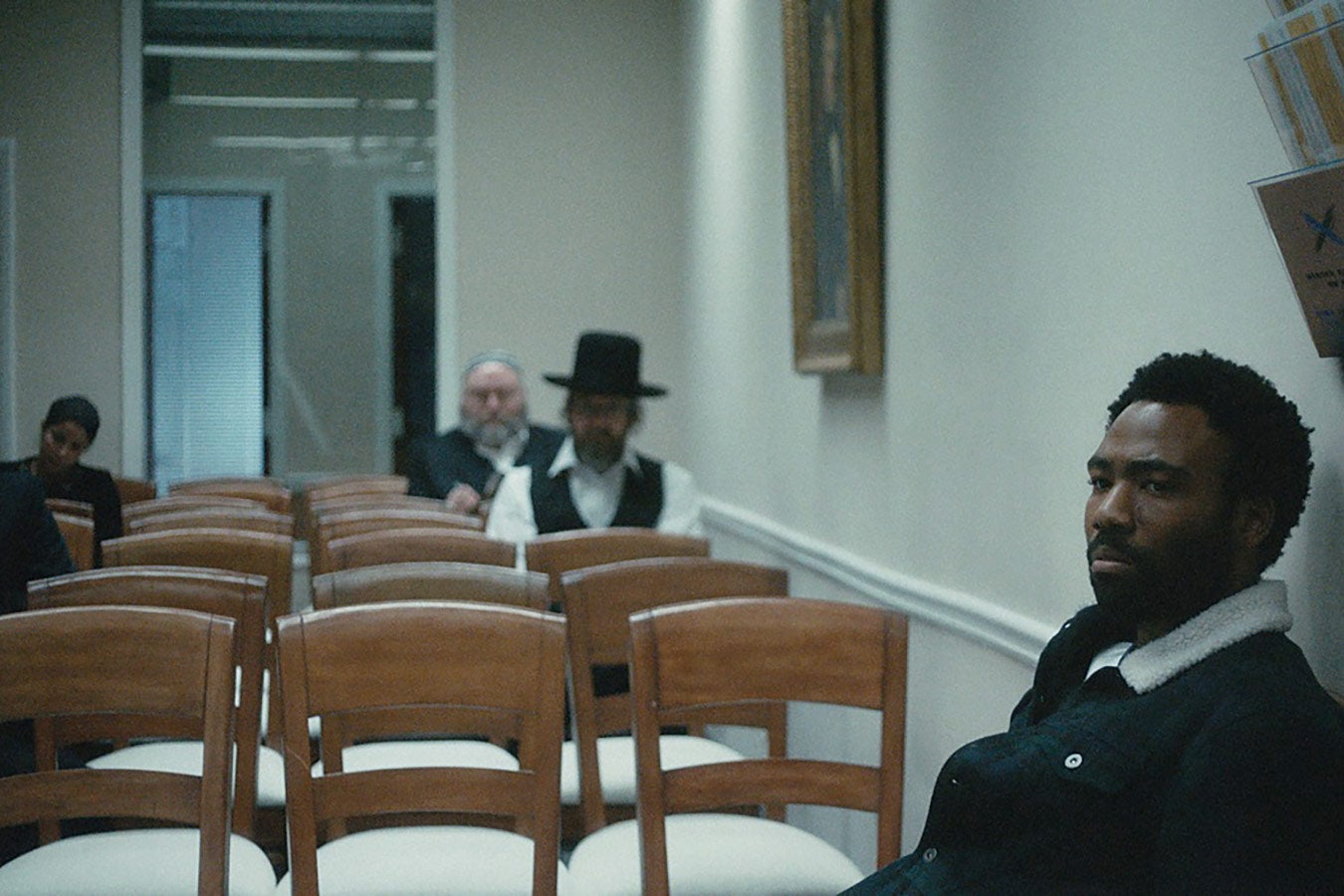 Donald Glover sits in a mostly empty waiting room looking pensive.