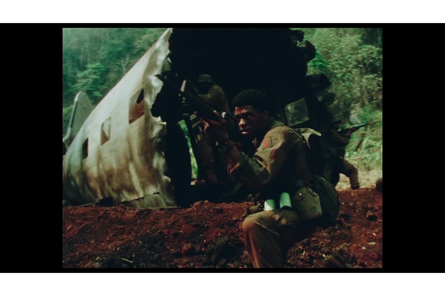 Chadwick Boseman holds up a rifle. Men in a destroyed airplane are seen behind him.