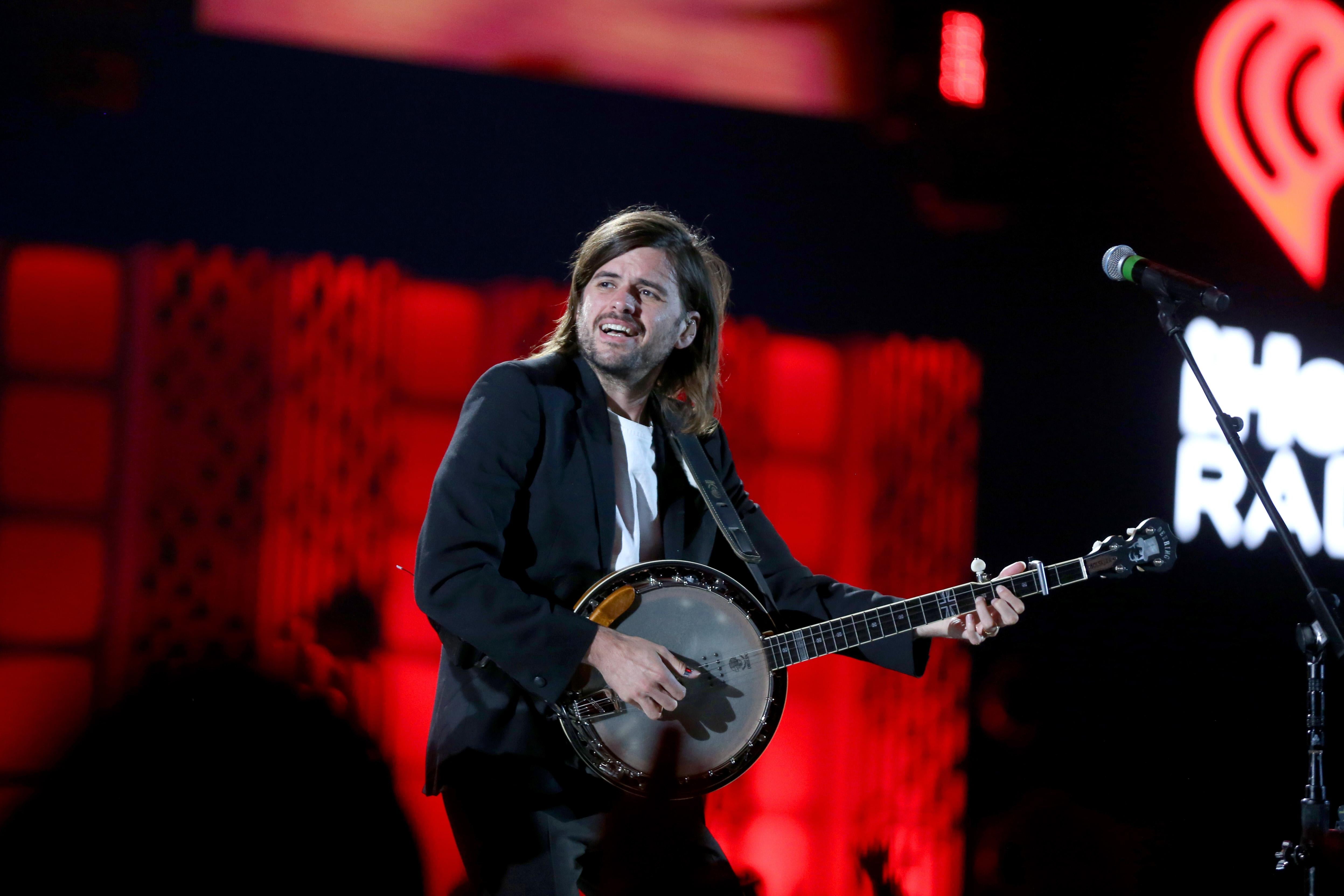 A man with long hair plays a banjo while wearing a black blazer and white T-shirt. He is on stage with a bright red light behind him. 