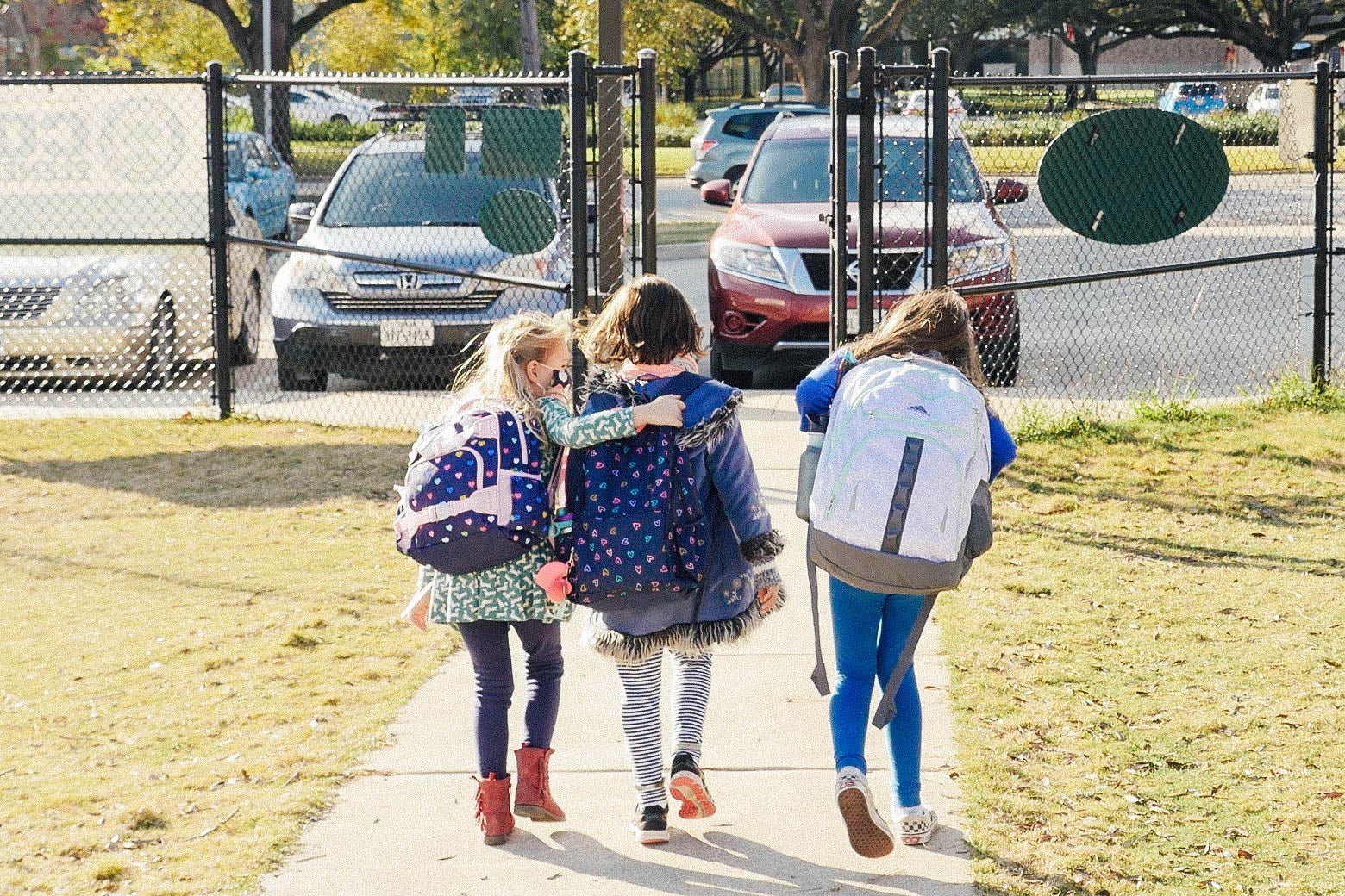 Three kids in backpacks walk down a sidewalk toward a parking lot.