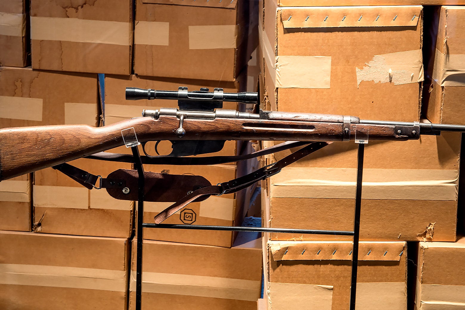 A wood-handled rifle with a shoulder strap and scope attached is seen before a wall of taped-up cardboard boxes. 