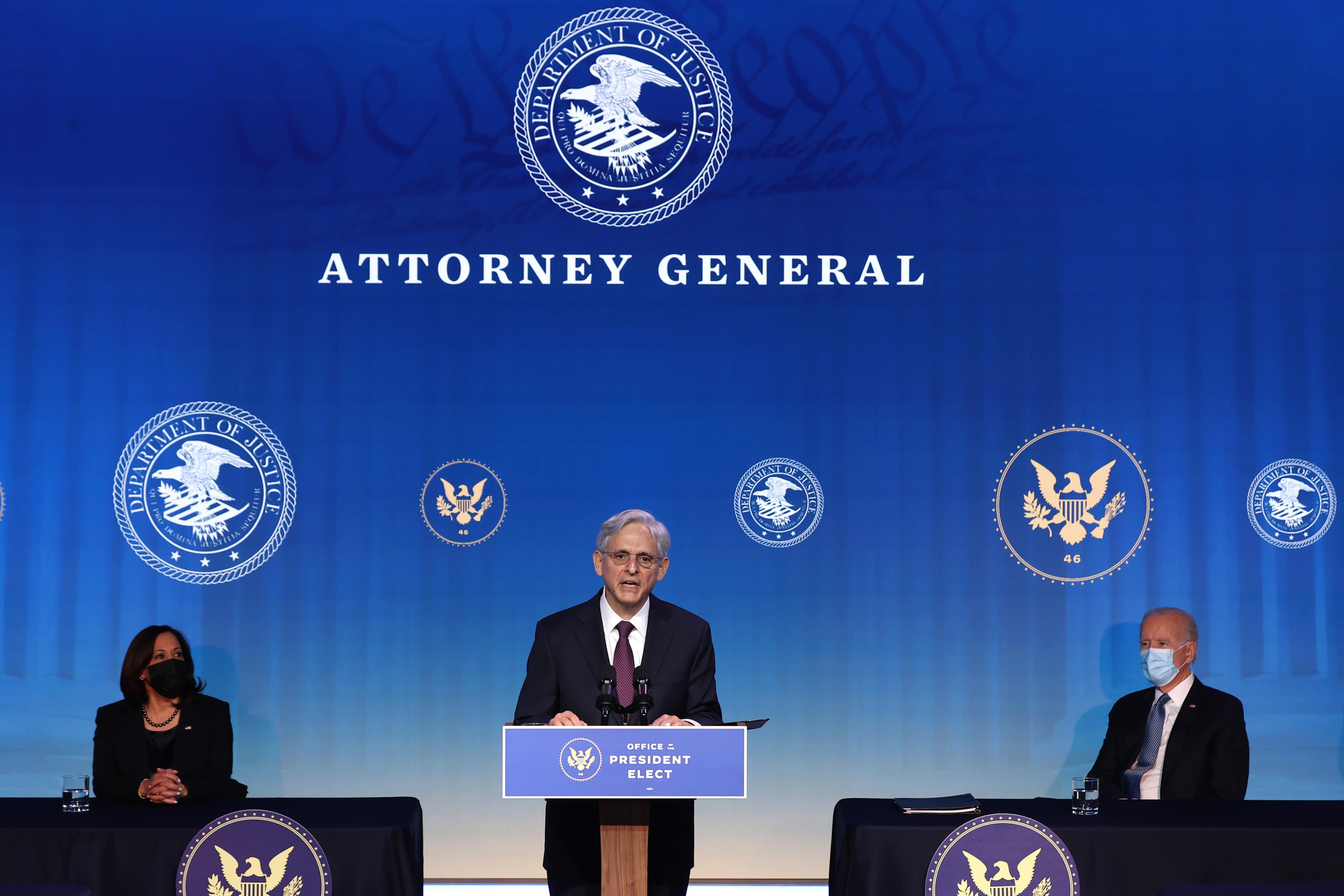 Merrick Garland speaks at a podium labeled Office of the President Elect as Joe Biden and Kamala Harris look on, seated on either side of him on a stage with a backdrop that says Attorney General.